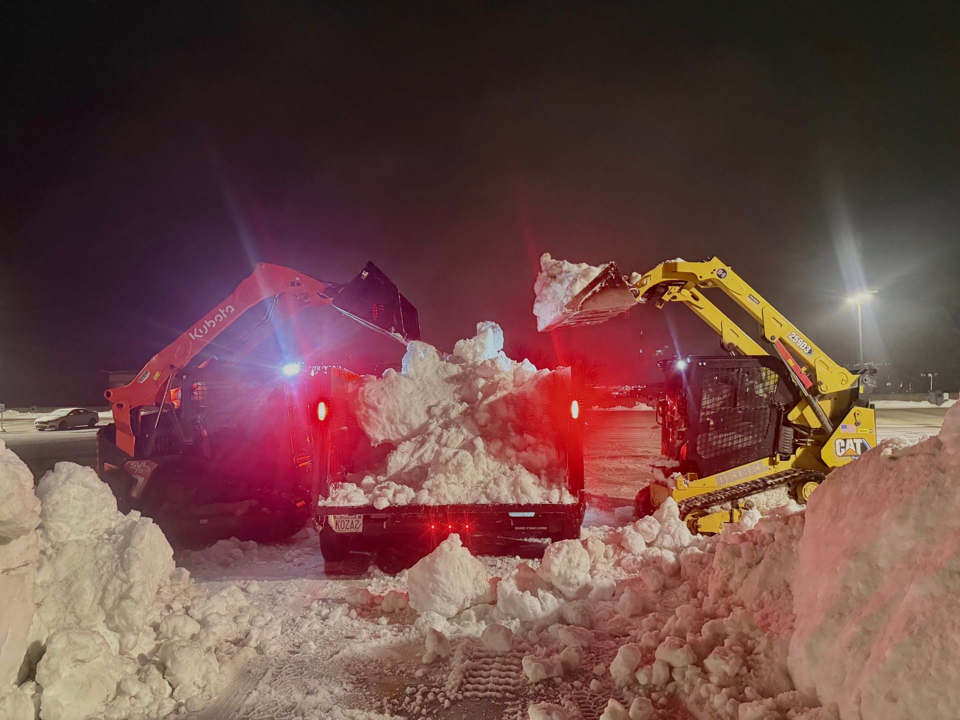 Two snow groomers clearing a snowy path at night, red and yellow lights glowing amid deep snowbanks