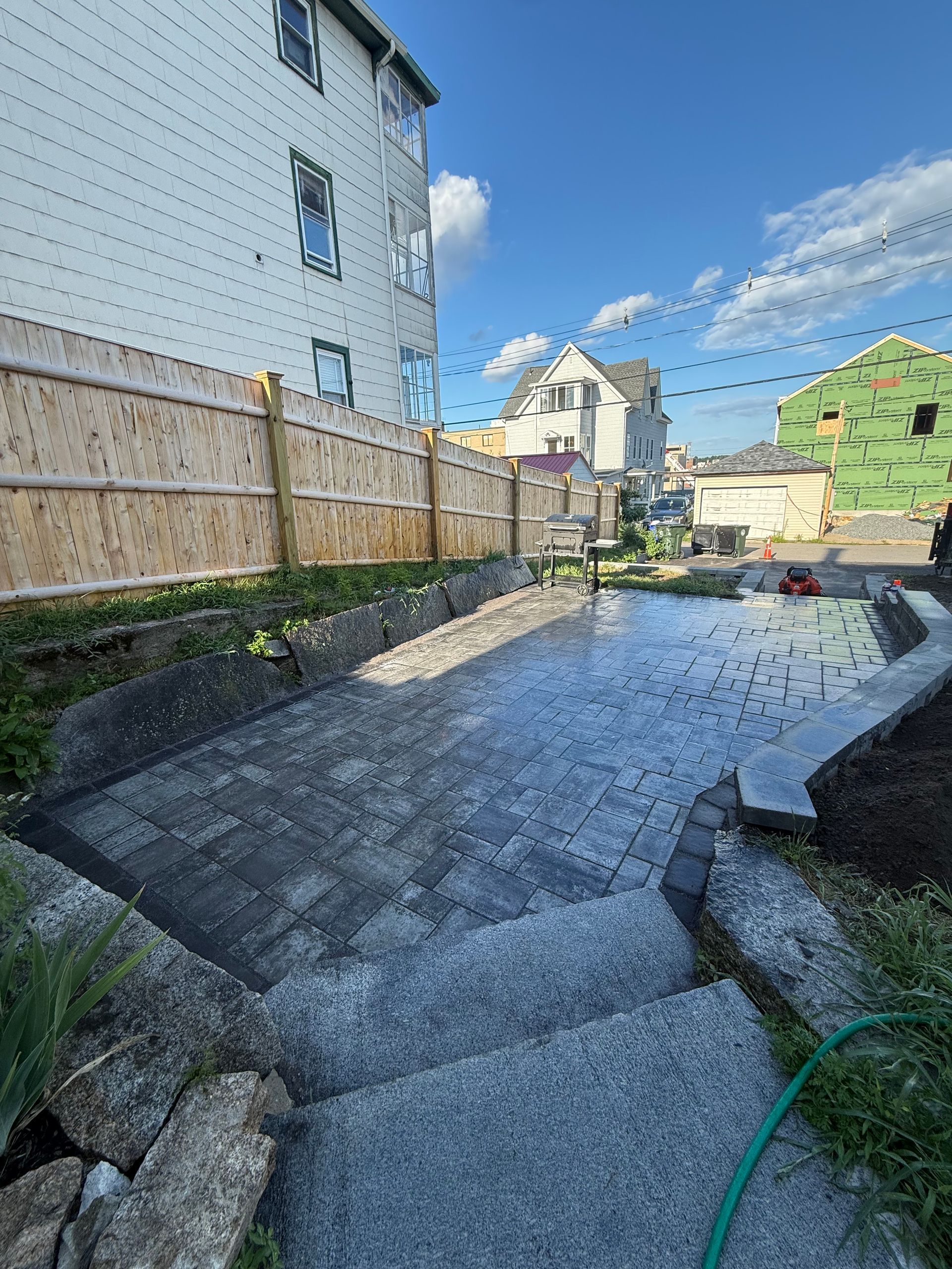 Concrete backyard walkway beside wooden fence and house, with neighboring homes under a blue sky.