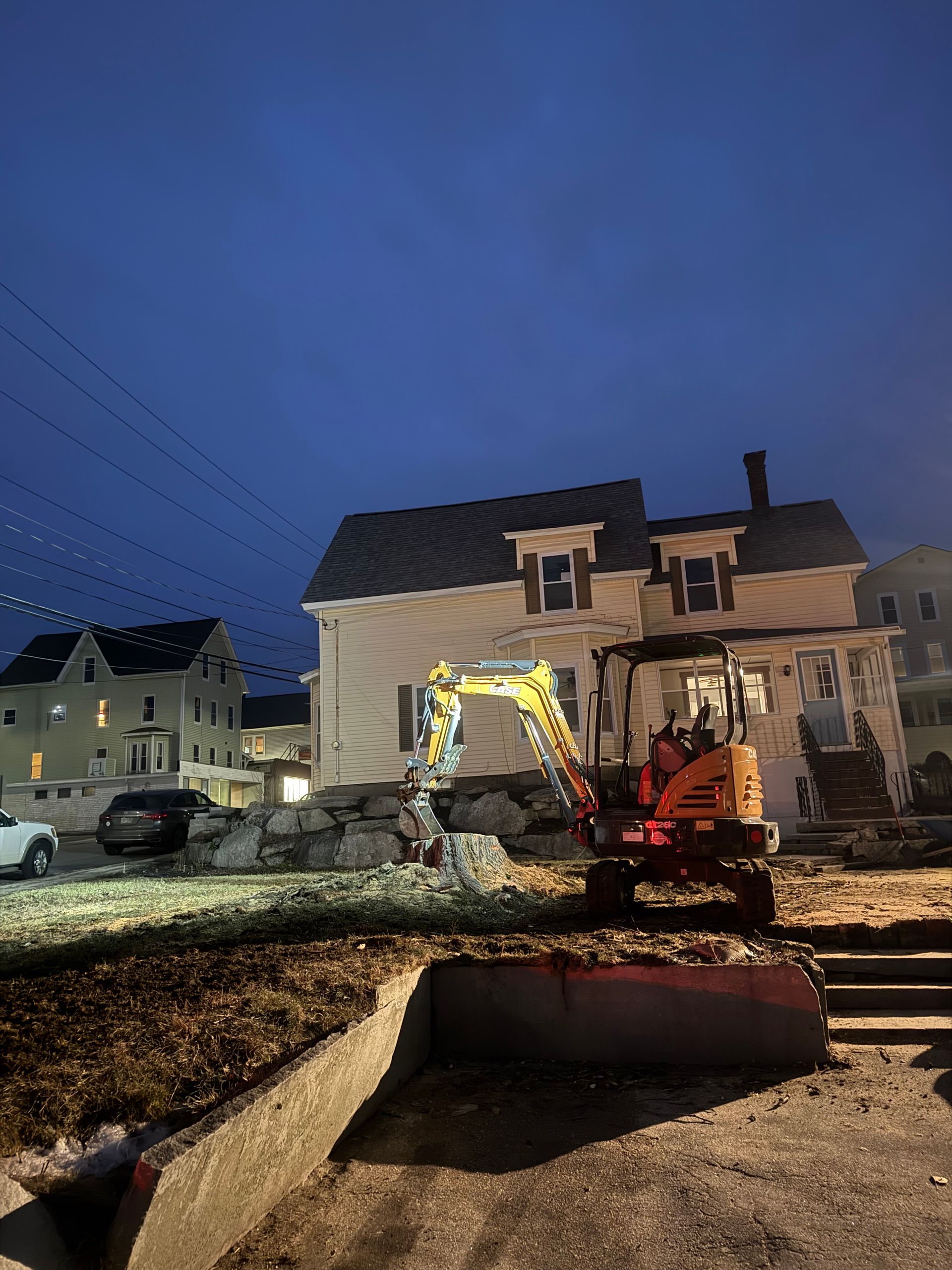 Excavator working on a front yard at dusk beside a two-story house, with dirt piles and construction equipment.
