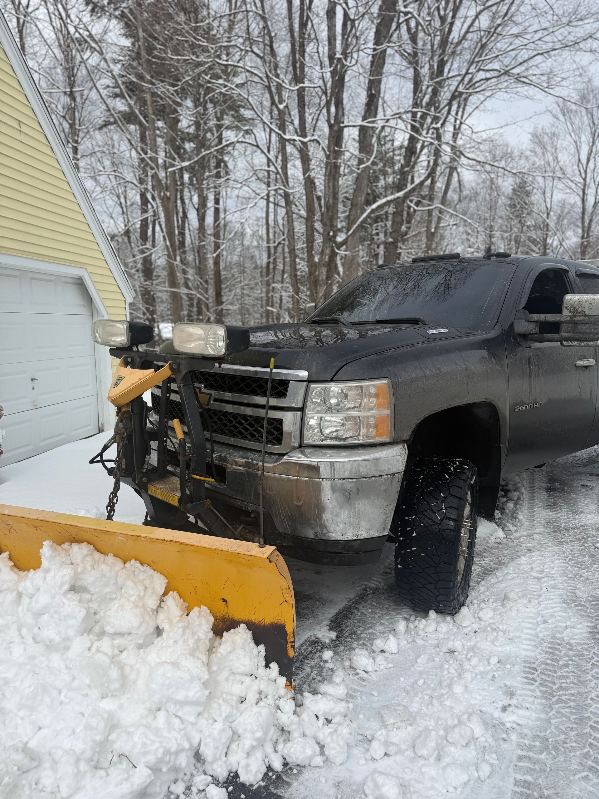 Black pickup truck clearing snow with front plow in a snowy driveway by a yellow house