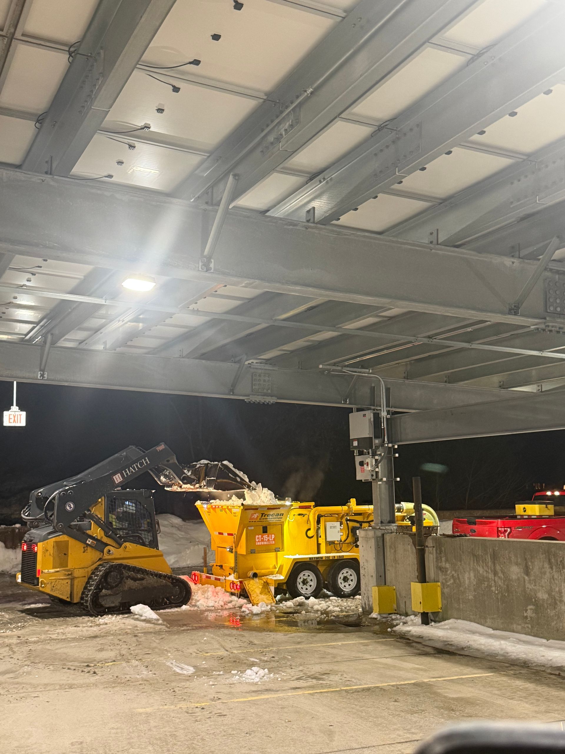 Yellow snowplow and dump truck under a covered work area at night, clearing snow near concrete barriers