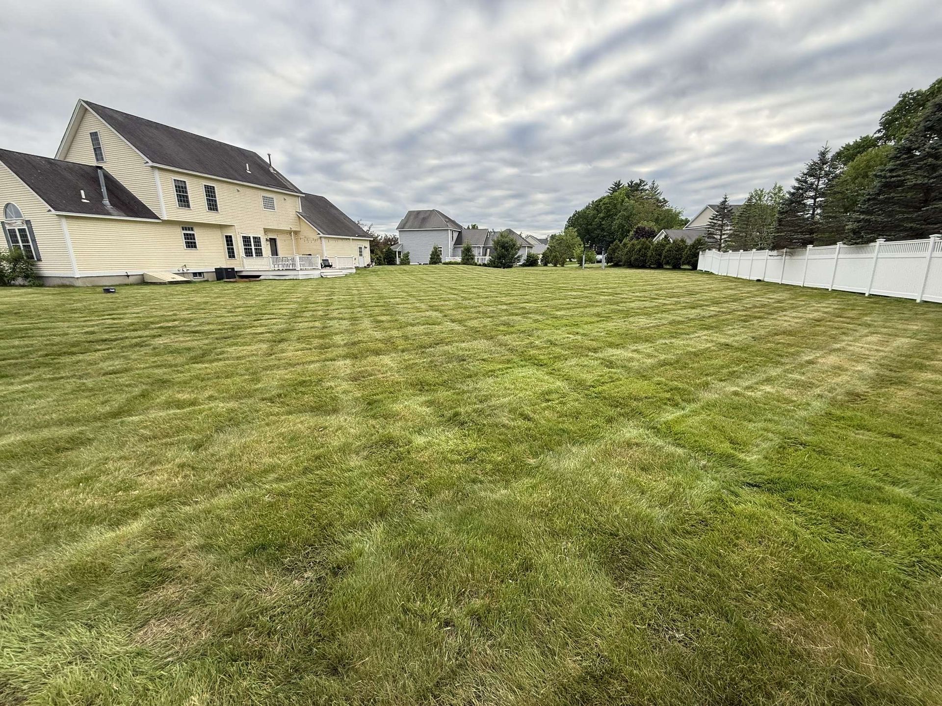 Wide grassy yard beside houses and a white fence under a cloudy sky.