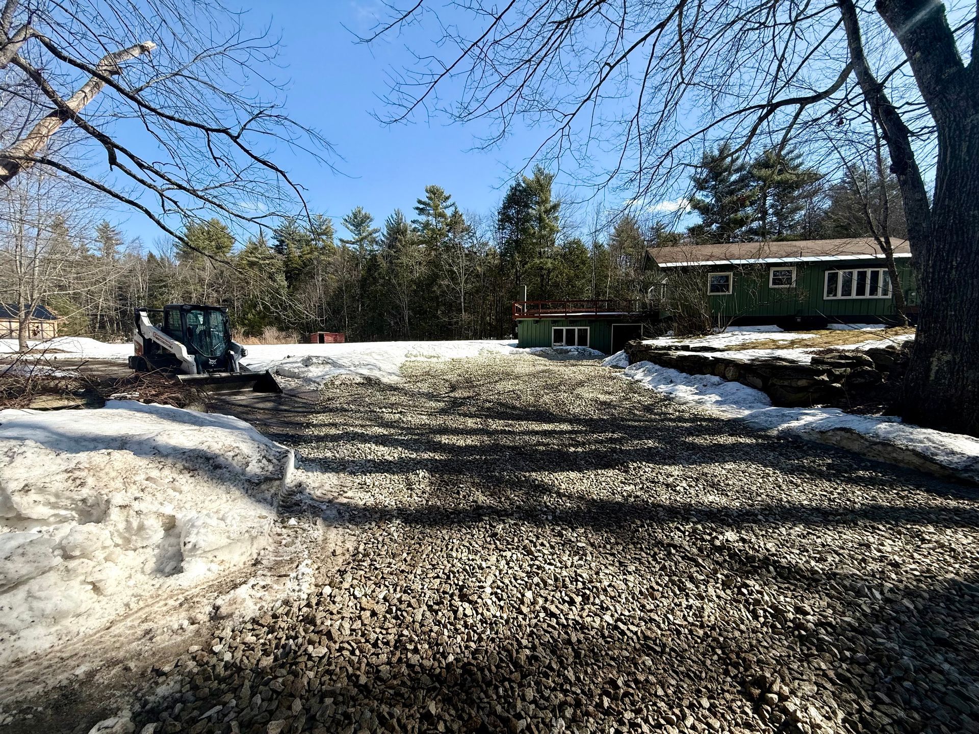 Snow-covered driveway beside a house, with trees casting shadows on a clear winter day.