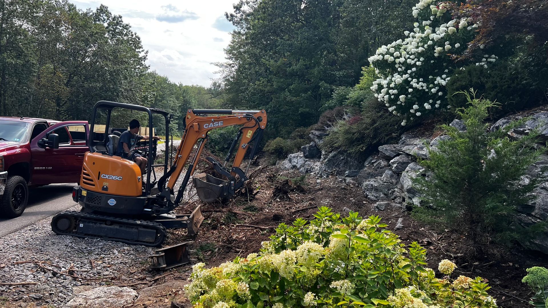 Orange excavator clearing rocks on a steep landscaped slope beside a red truck and trees