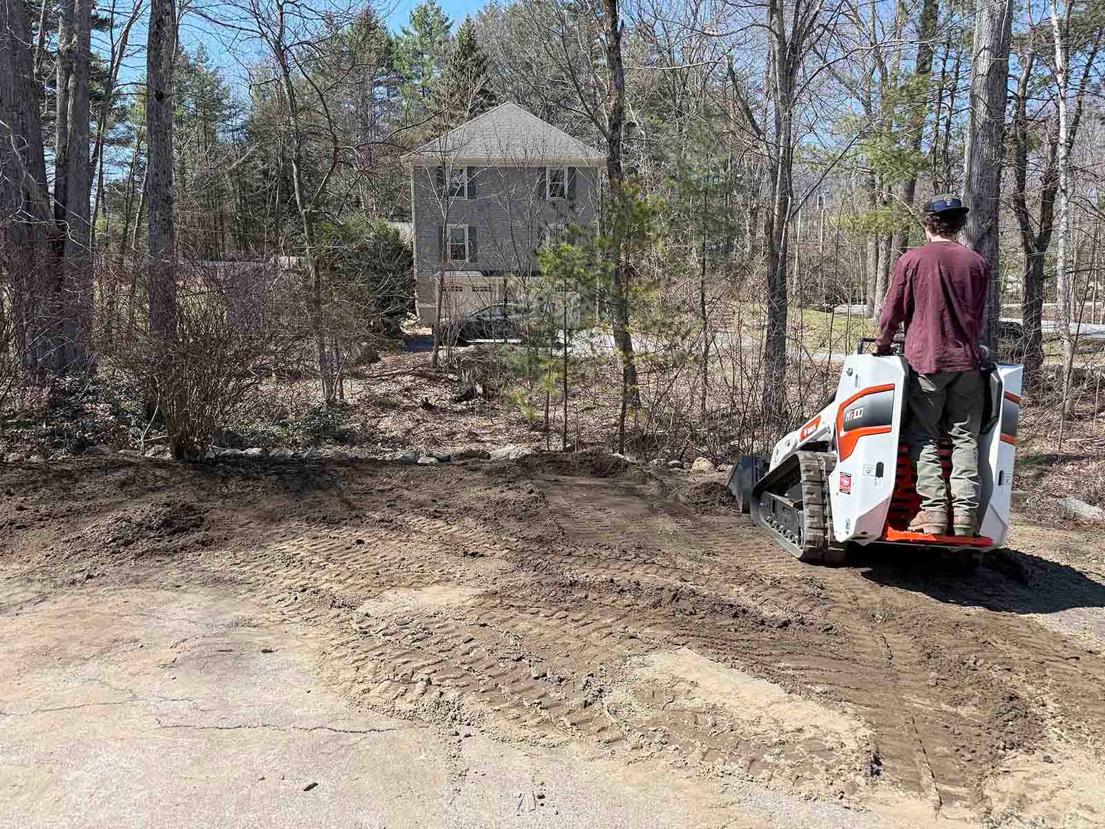 Person operating a skid steer on a dirt driveway beside a wooded lot and house under construction