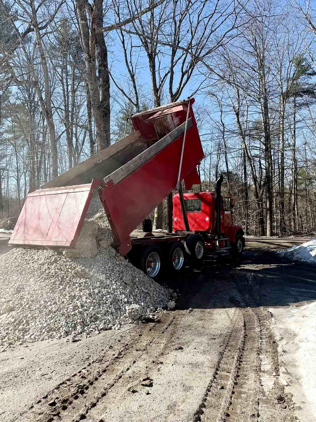 Red dump truck tipping out a load of gravel onto a muddy forest road.