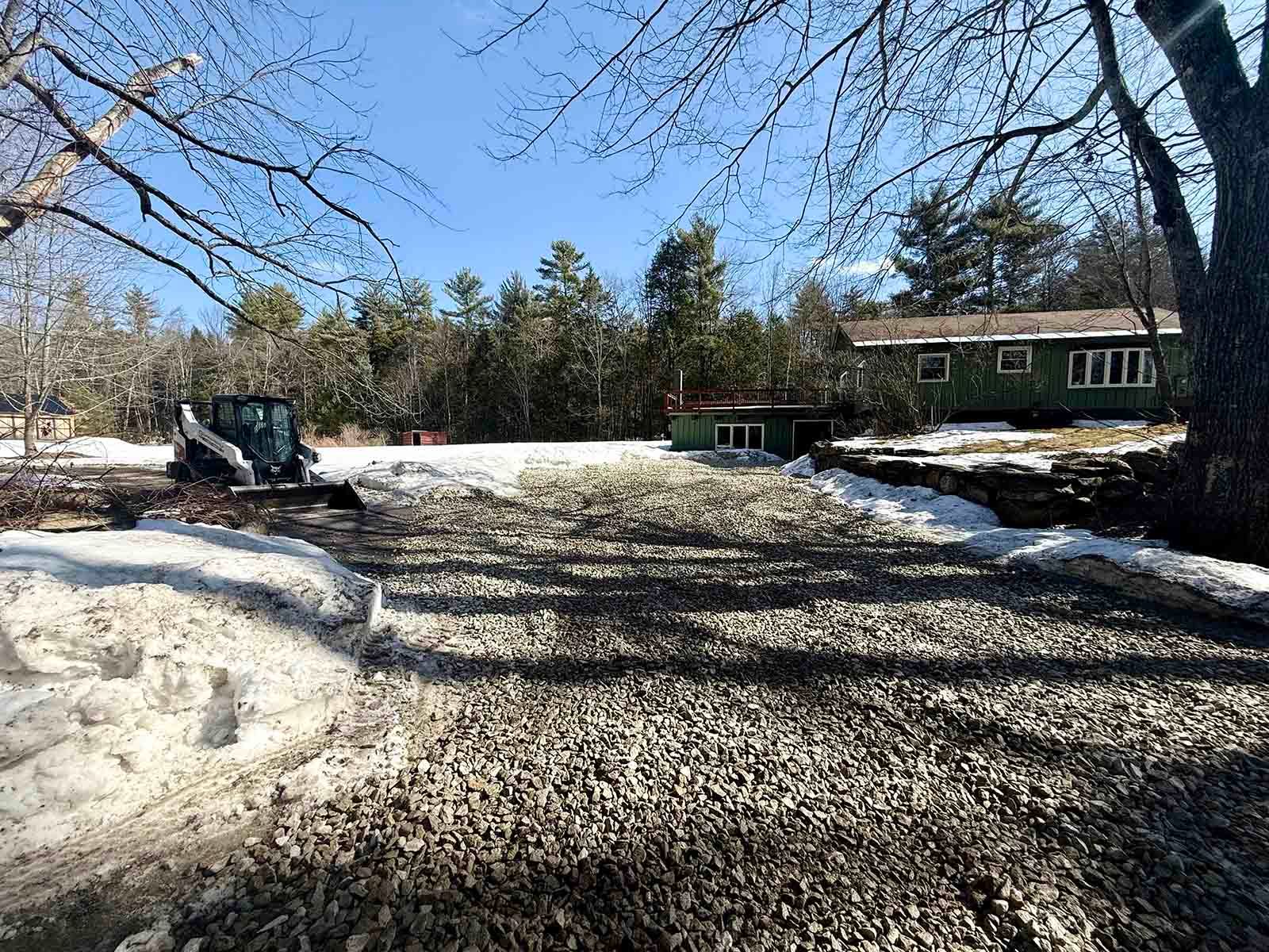 Snow-covered driveway with a parked excavator and a house under a clear blue sky