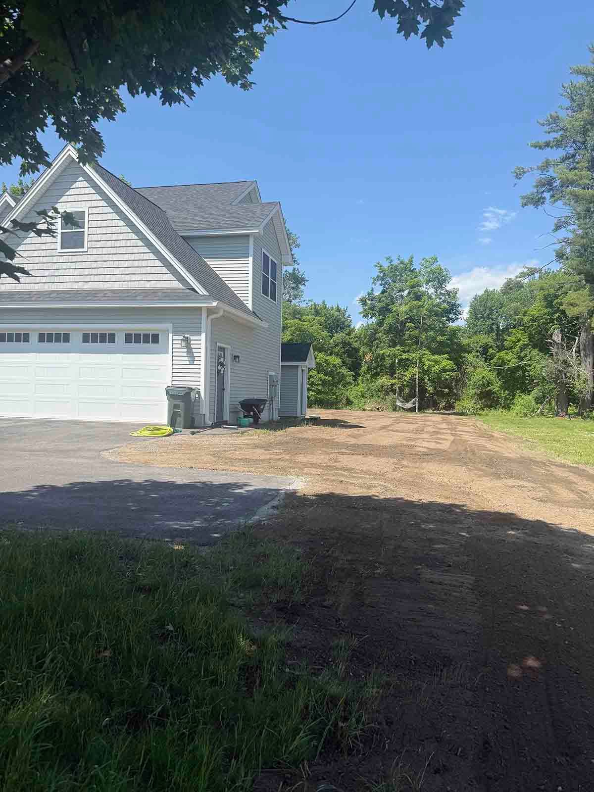 White house with garage on a dirt driveway beside trees and open yard under a clear blue sky.