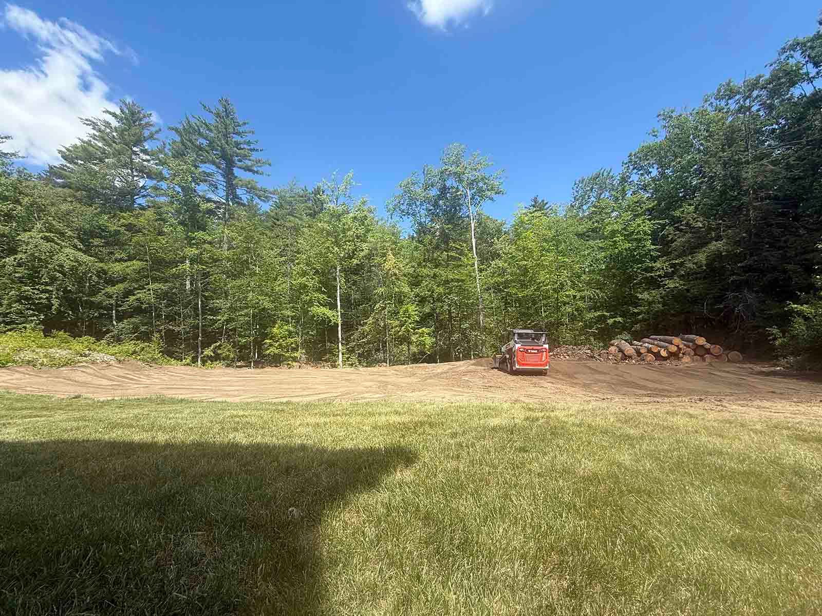 Grassy clearing beside a tree line under a blue sky, with a red mower or utility vehicle near the woods.