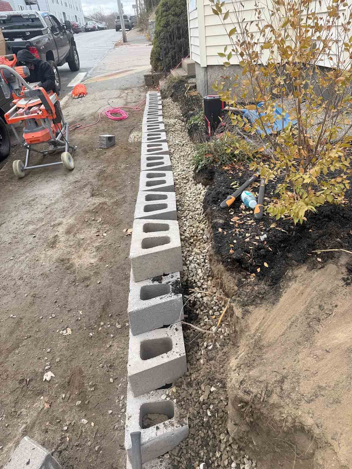 Row of concrete cinder blocks lining a dirt trench beside a sidewalk and house under construction.