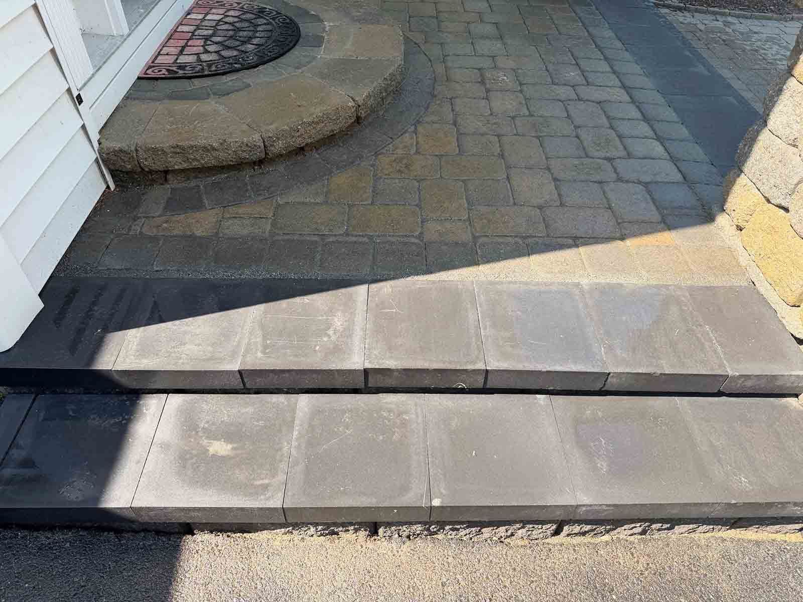 Low curb ramp from sidewalk to paved path beside a white building, with shadowed steps and pavers.