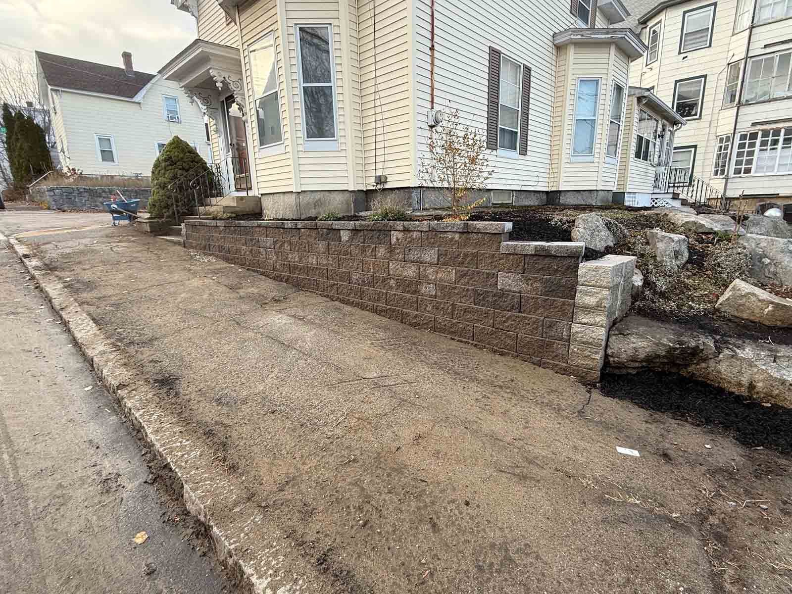 Sloped concrete driveway beside a white house, with a brick retaining wall and stone steps.