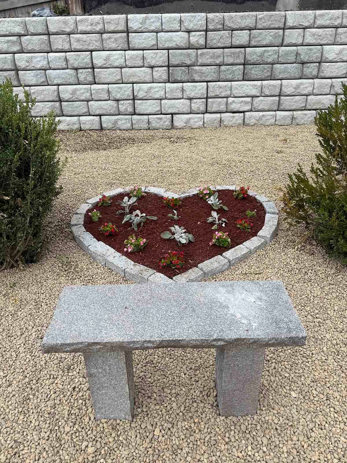 Heart-shaped flower bed with red mulch and small plants behind a stone bench and low brick wall.