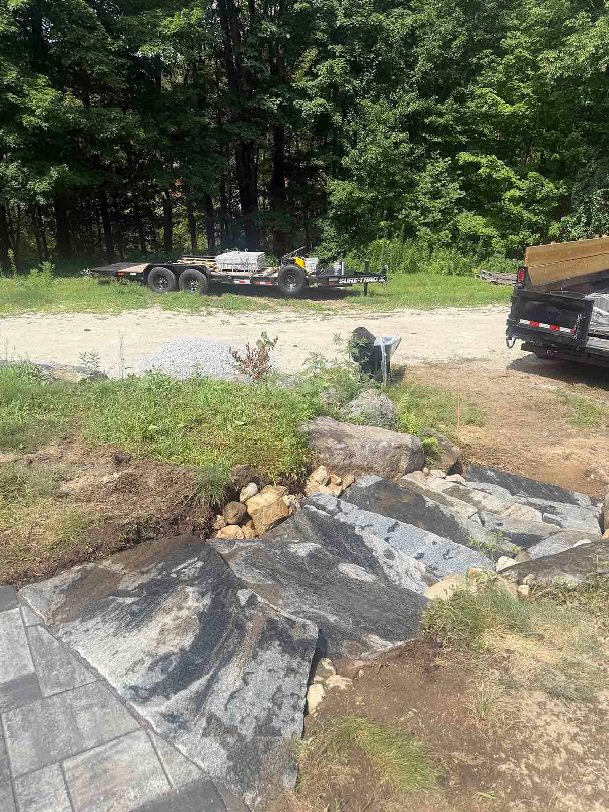 Rocky patio and gravel driveway with parked cars beside a wooded yard