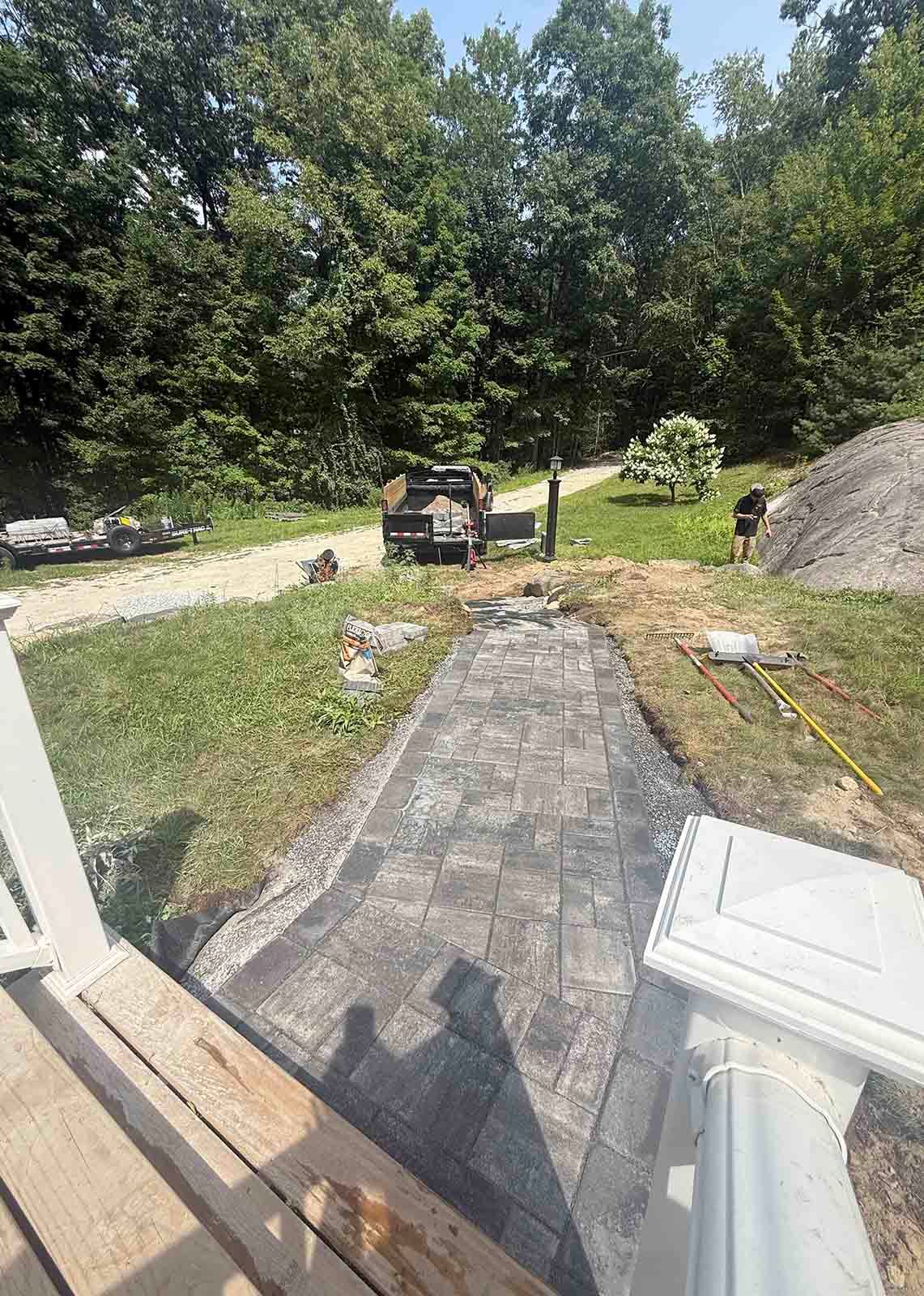 Stone patio leading to a grassy yard with trees, a grill, and a large rock in the background.