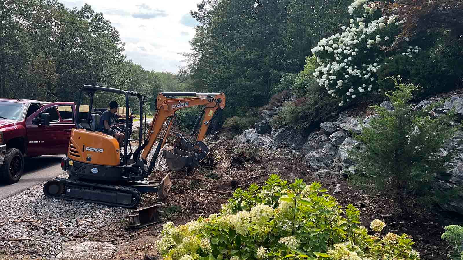 Orange excavator clearing brush beside a red truck on a rocky hillside under cloudy sky
