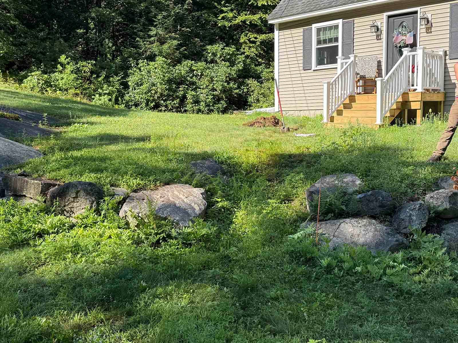 Small house with stairs on a grassy hillside, bordered by rocks and lush green trees.