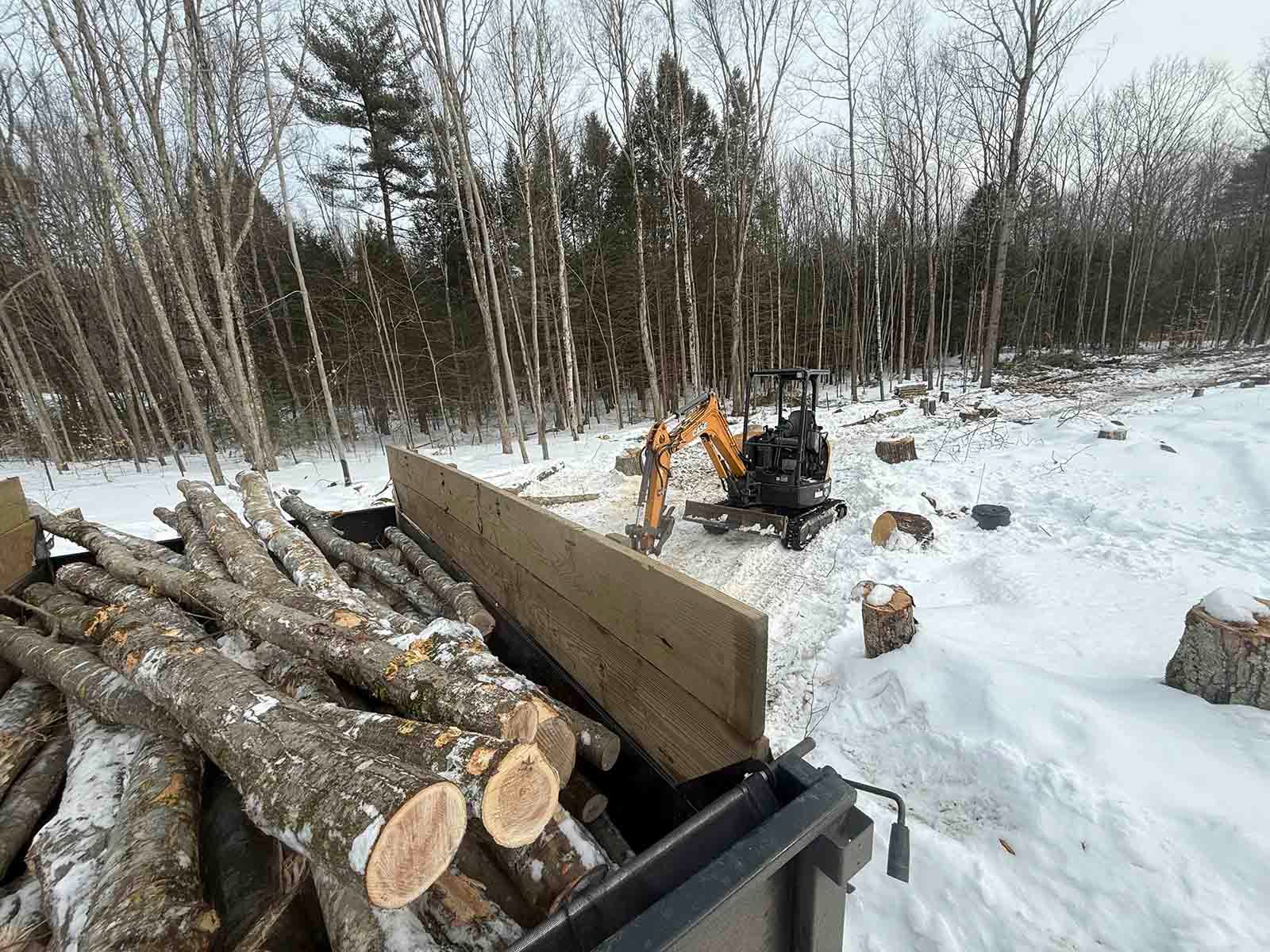 Logs loaded onto a trailer in a snowy wooded area with an excavator nearby