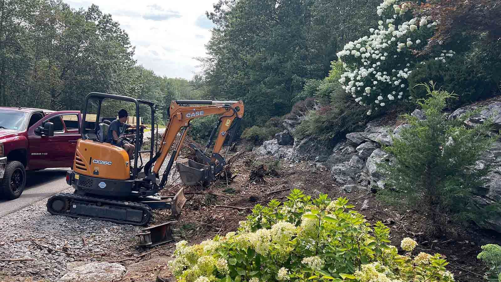 Yellow excavator clearing debris beside a red truck on a wooded rocky roadside
