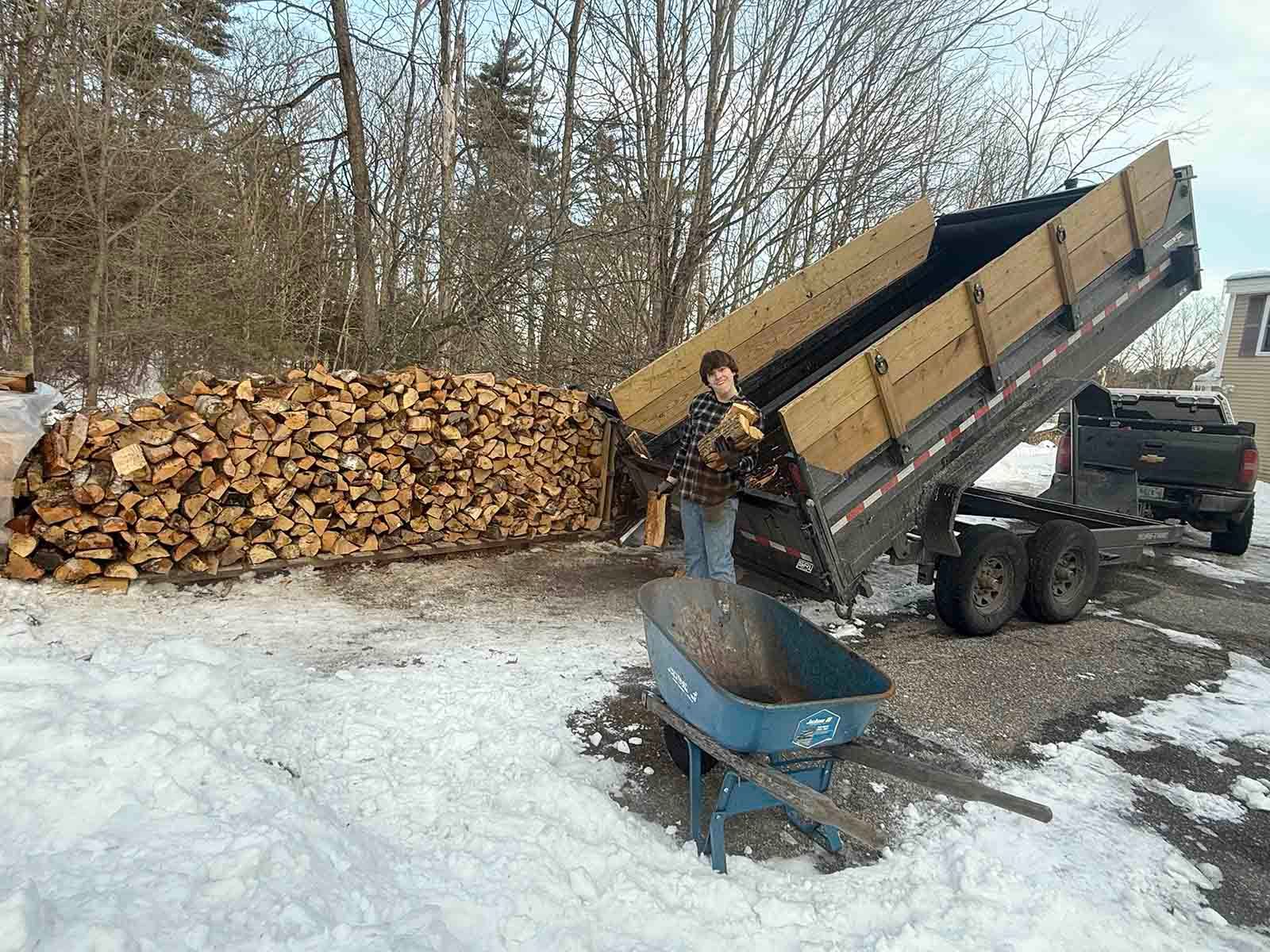 Dump truck tipping a load of firewood into a snowy yard beside a large stack of logs