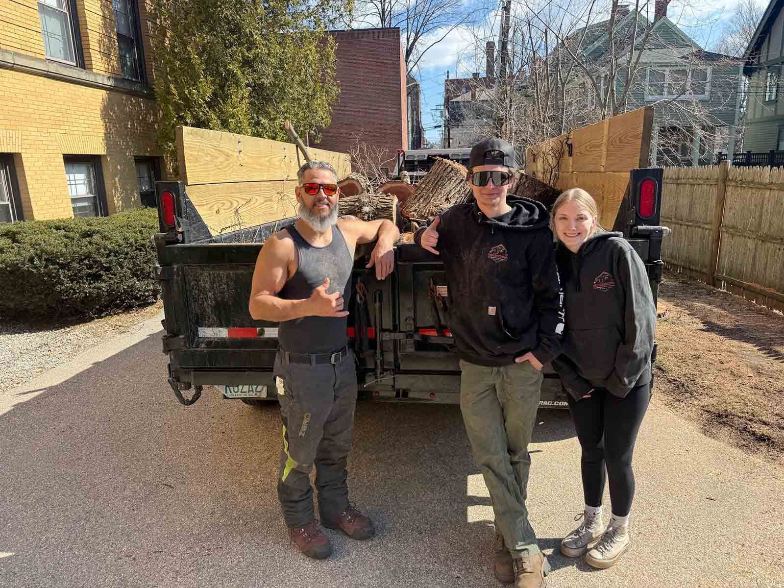 Three people posing beside a black truck in a sunny driveway.