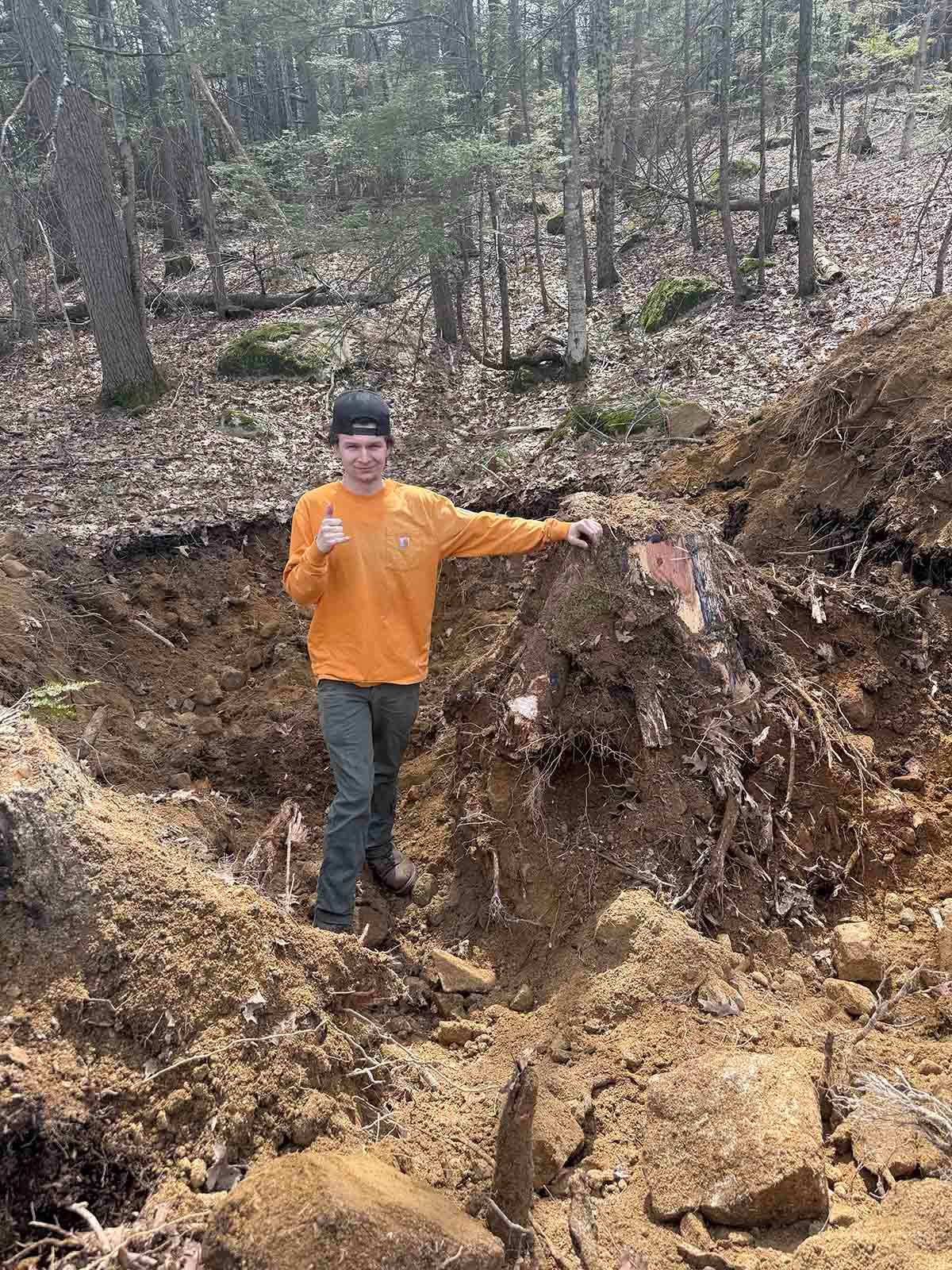 Person in orange shirt standing in a muddy forest excavation site, holding out one arm.