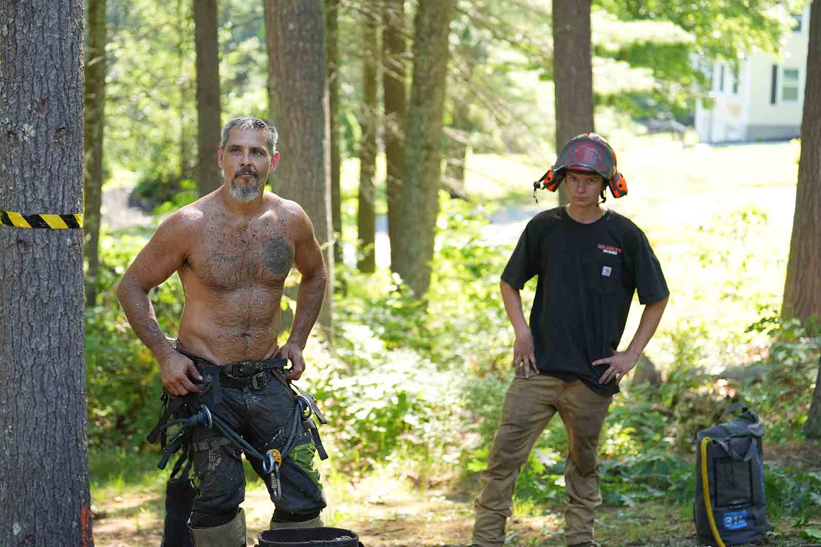 Two workers standing in a wooded area, one shirtless, both wearing work gear near a tree and equipment