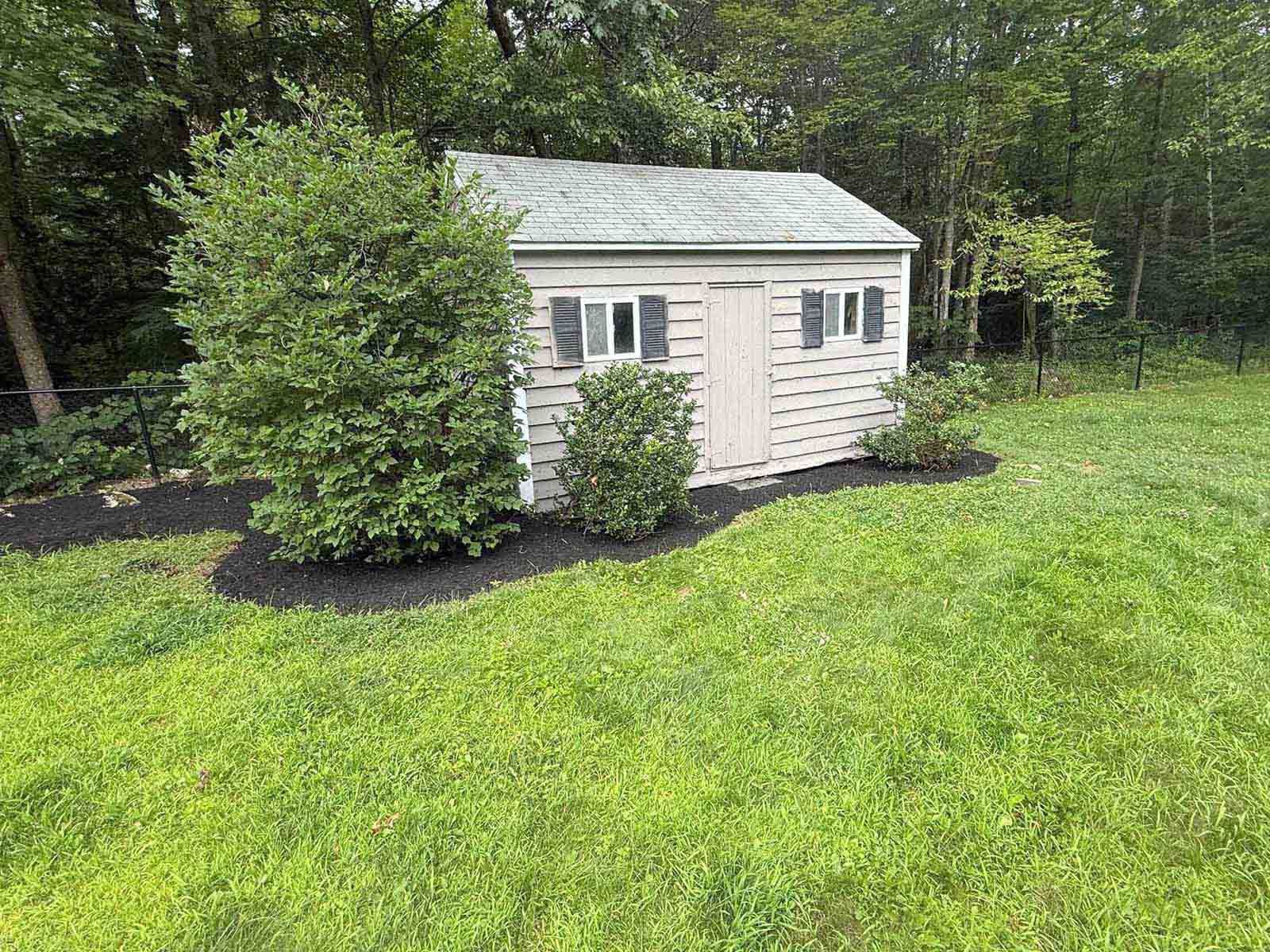 Small beige garden shed with white trim beside bushes and lawn in a wooded yard