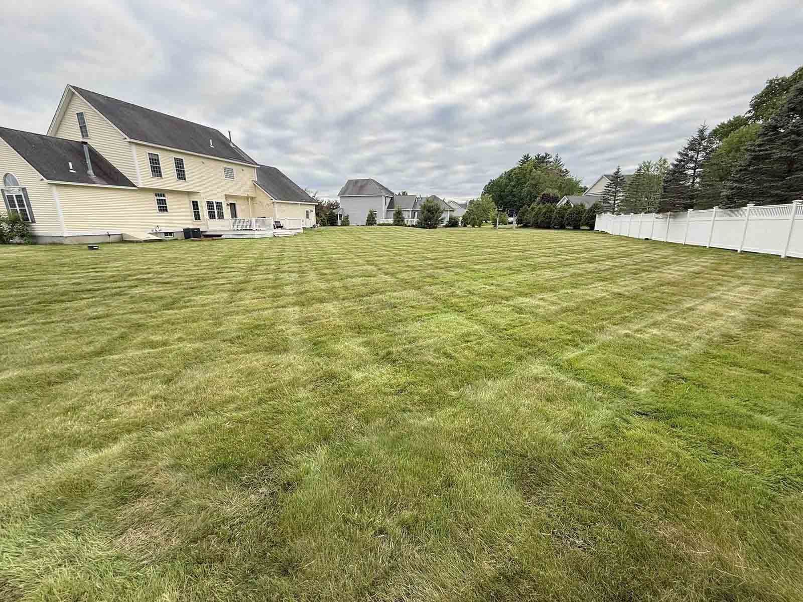 Wide grassy yard beside a white house and fence under a cloudy sky