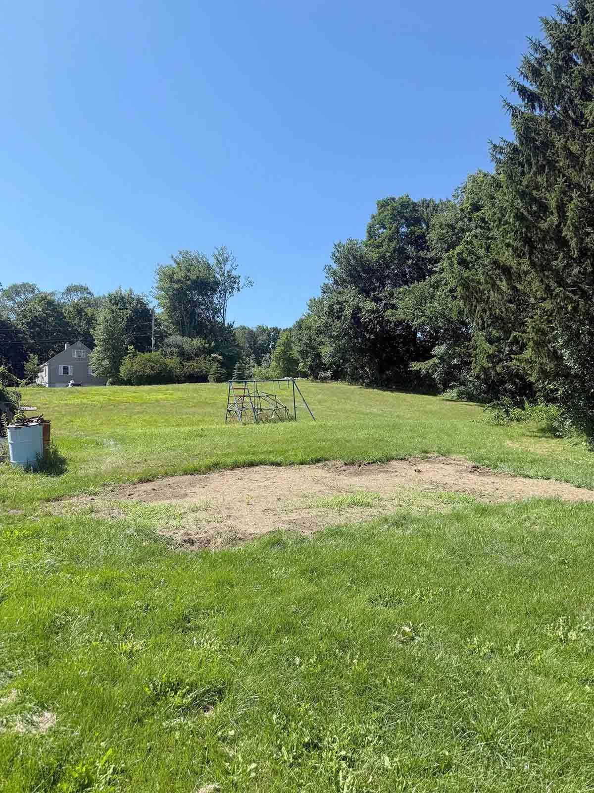 Sunny grassy yard with a dirt patch, trees, and a small garden chair under a blue sky
