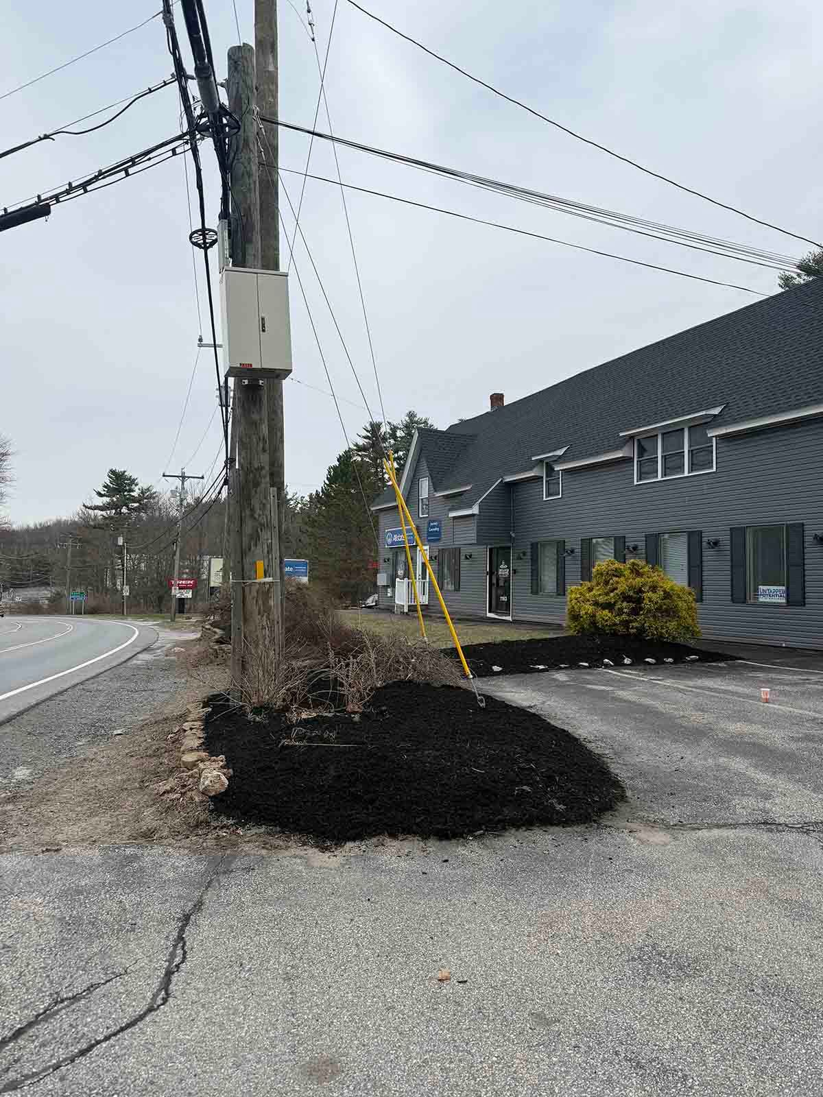 Roadside utility pole beside a gray house, with a mound of dark soil and utility lines overhead