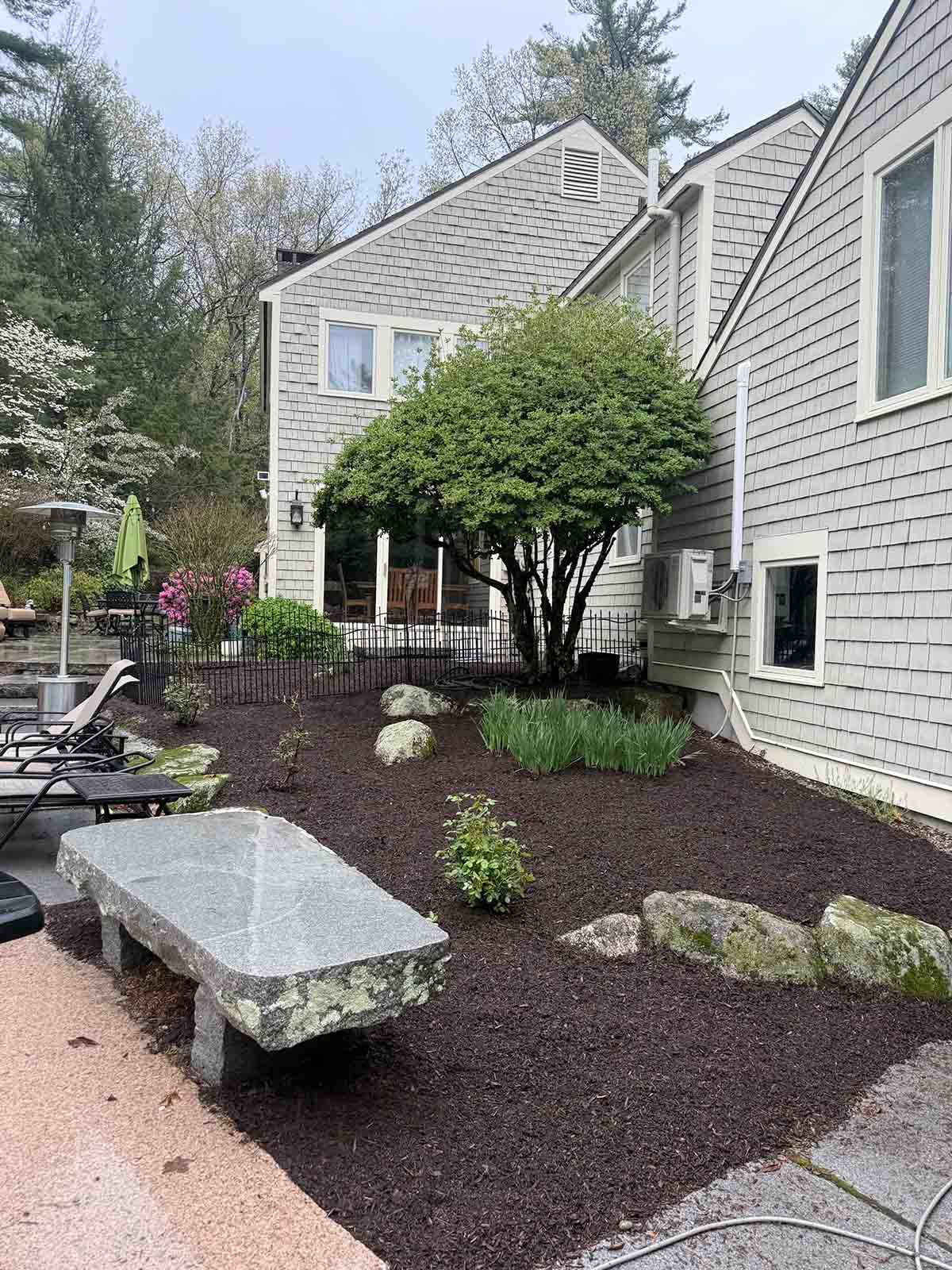 Landscaped side yard with mulch beds, shrubs, stepping stones, and a stone bench beside a gray house