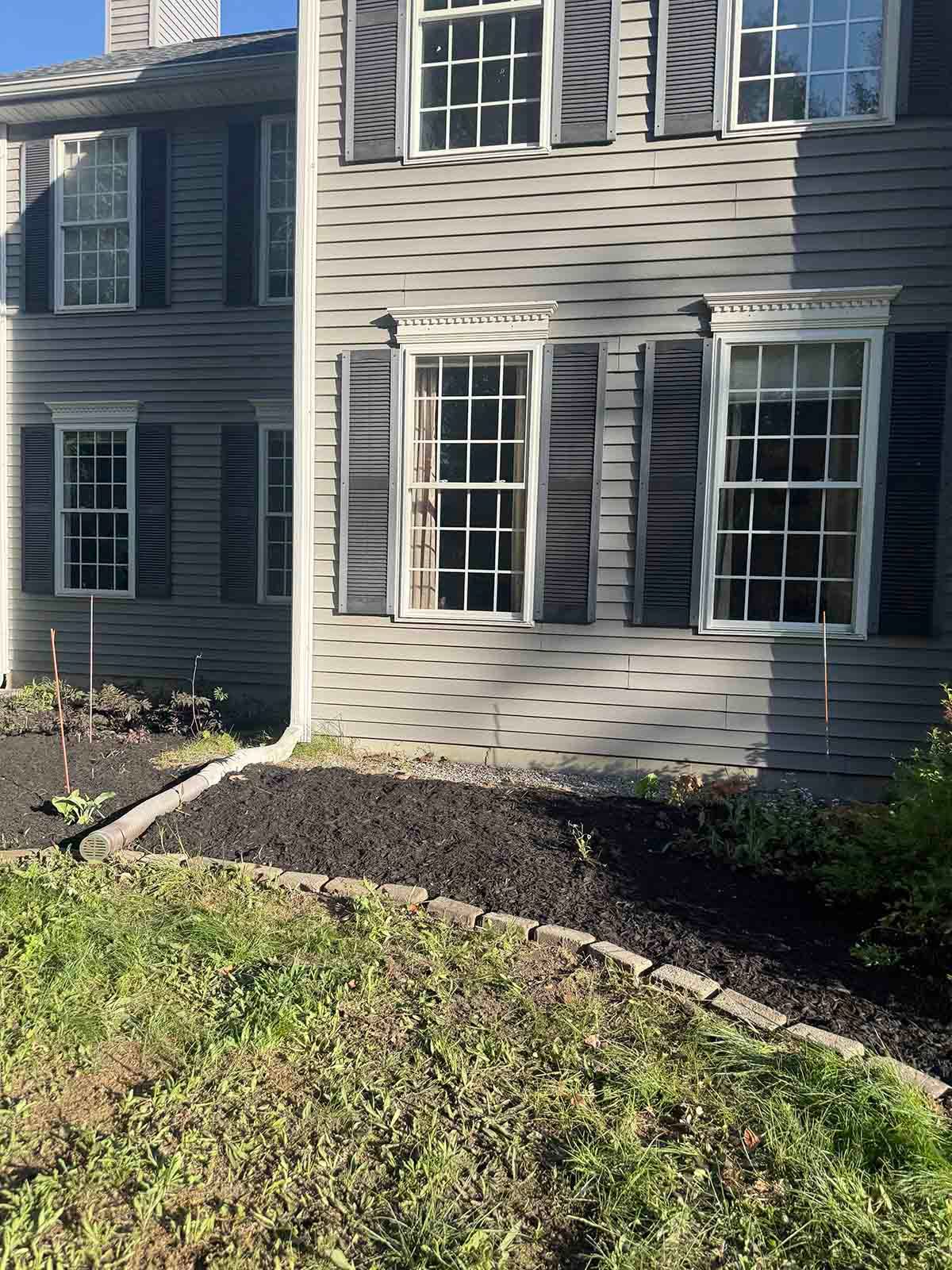 Gray house exterior with white-trimmed windows, stone border, and mulch bed along the front lawn
