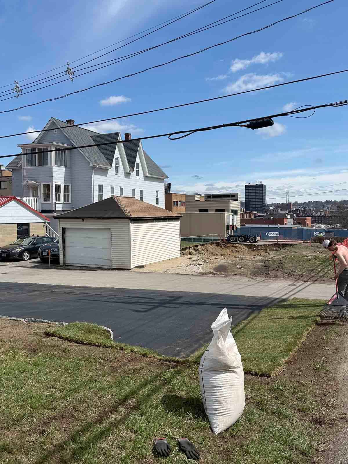 Street with house, utility lines, and a small shed by a waterfront under a clear blue sky.