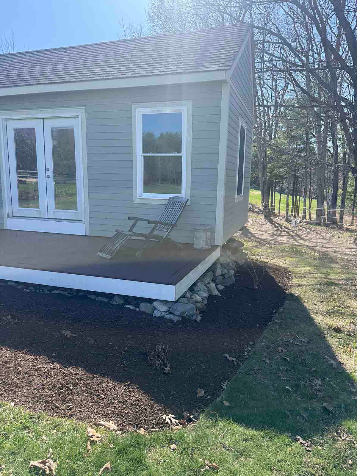 Small gray house with white trim, double doors, a window, and a porch chair beside a wooded yard.