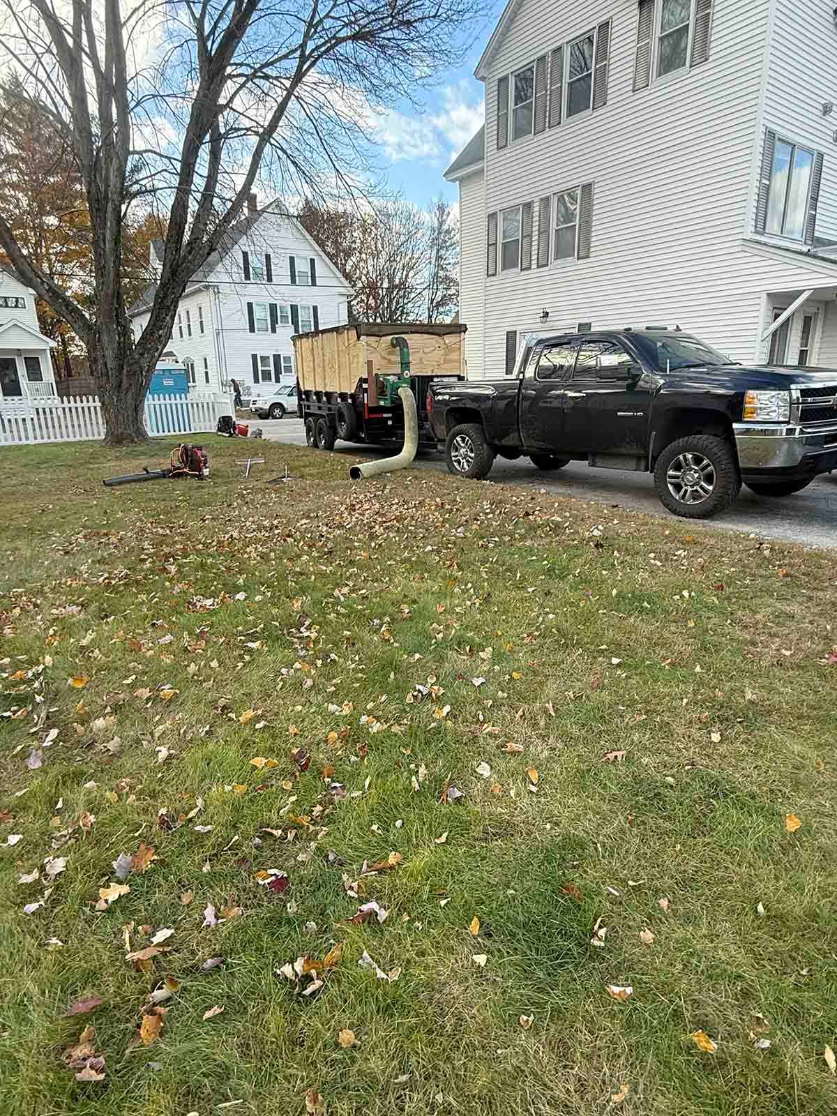Black pickup truck parked beside white houses on a grassy yard with fallen leaves, people in the background