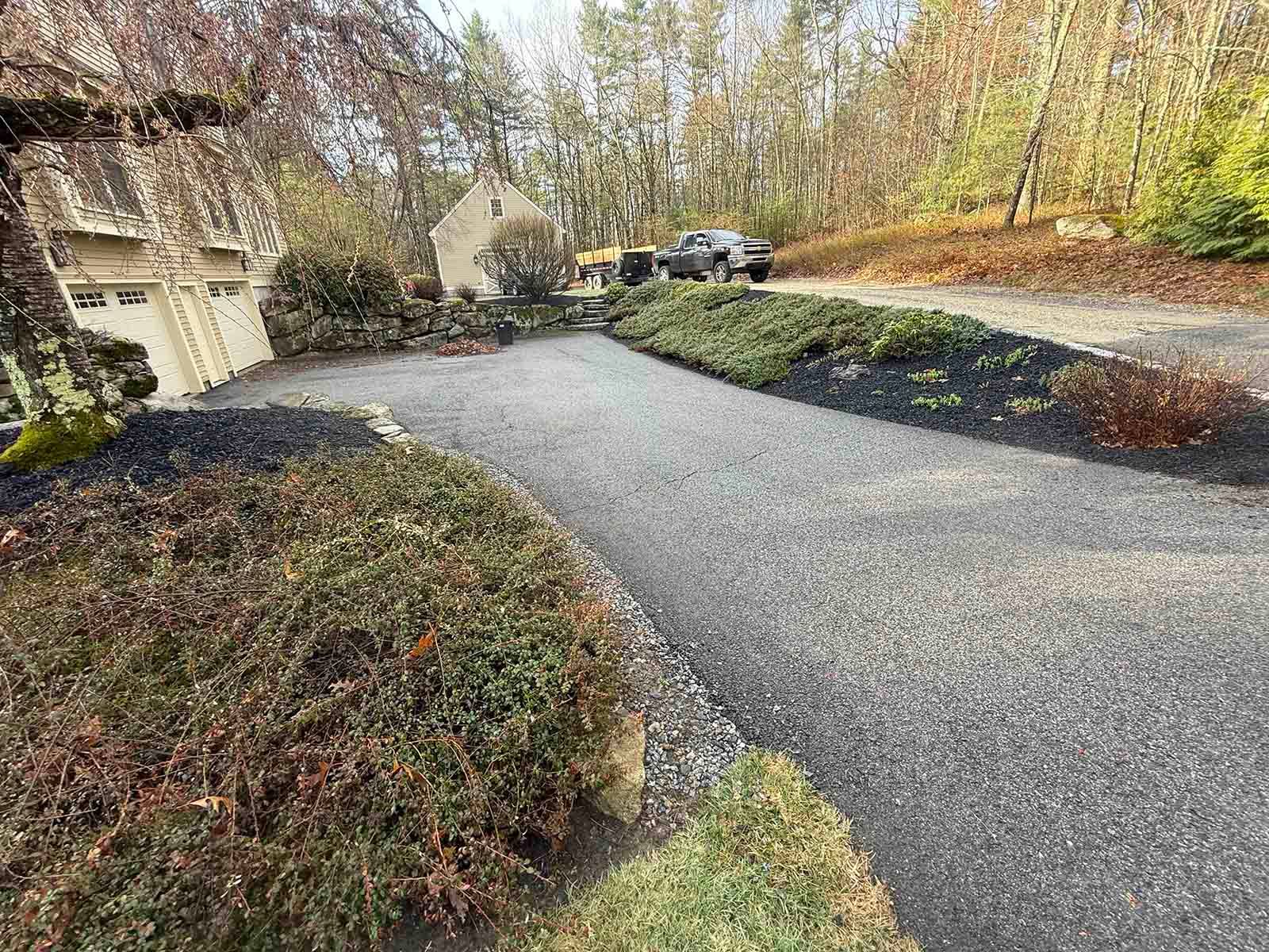 Curved gravel driveway beside a house, with bushes and trees in a wooded yard