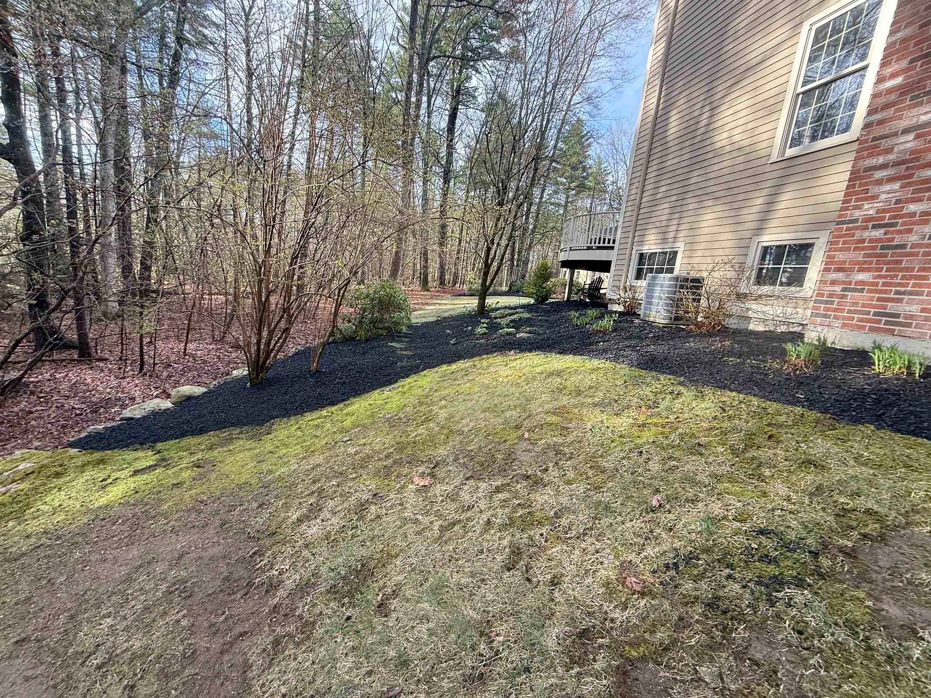 Side of a brick house with a sloping grassy yard and mulched border beside leafless trees