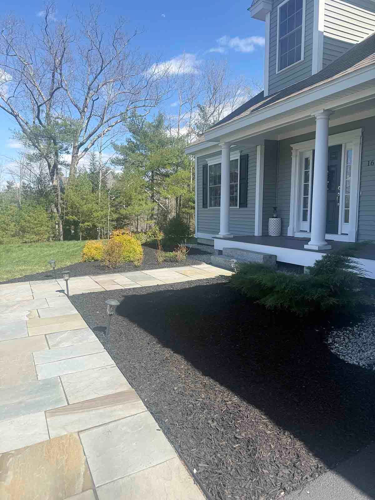 Front porch with white columns beside a stone walkway, bordered by mulch and a grassy yard under a blue sky.