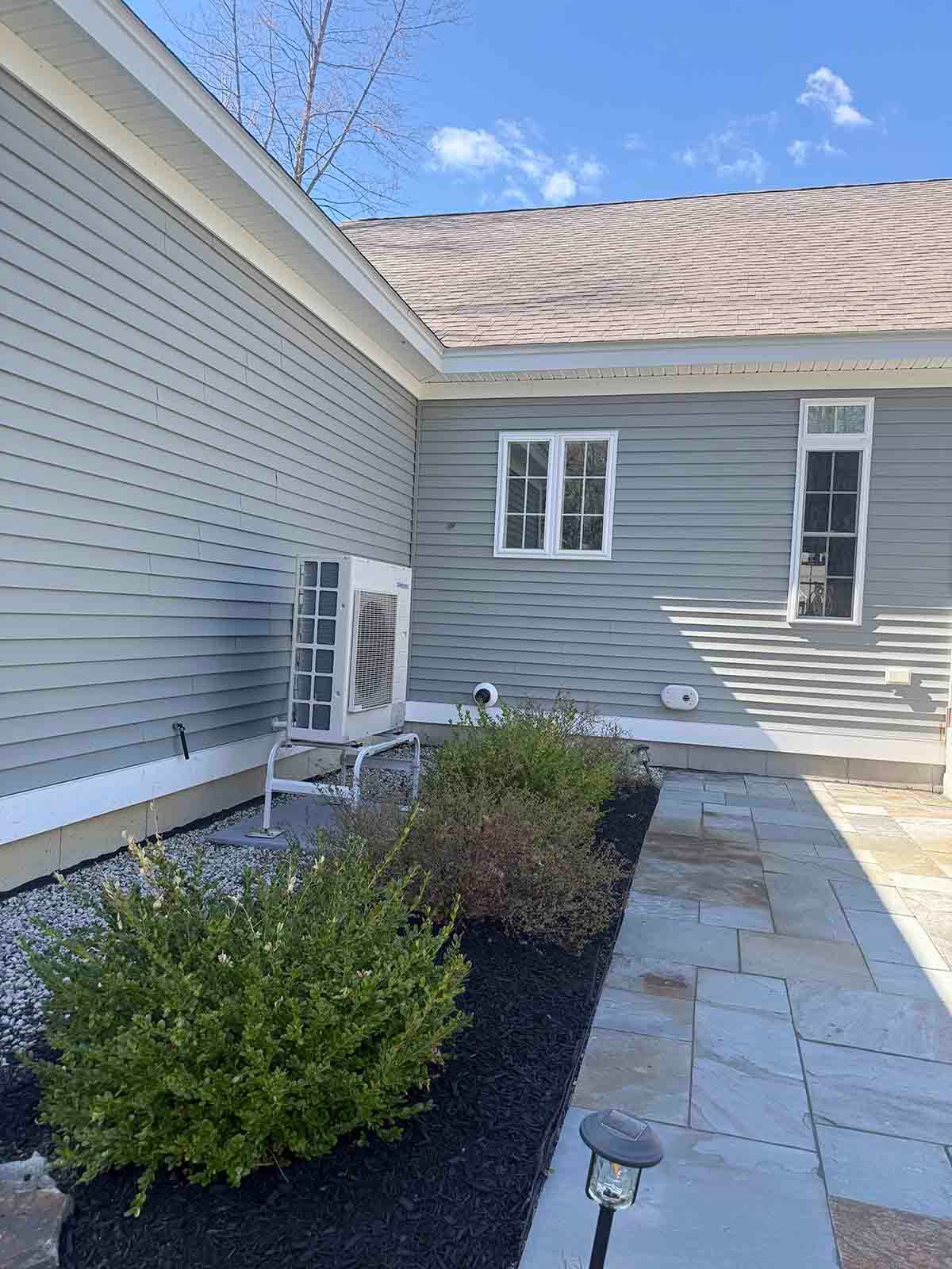 Gray-sided house exterior with stone patio, shrubs, and a small garden light under a blue sky