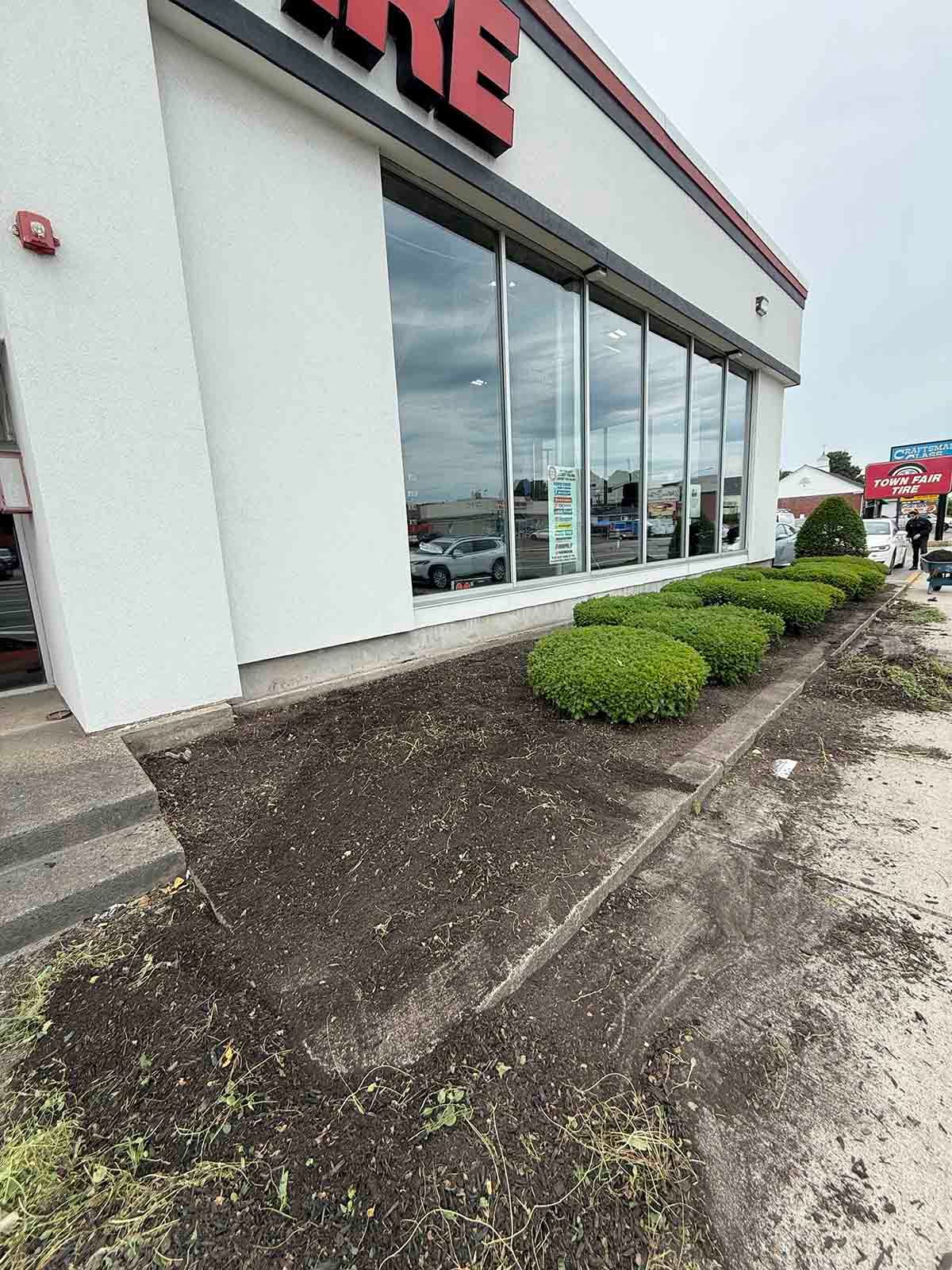 Exterior of a store with large windows, white facade, and a muddy, sloped walkway with shrubs