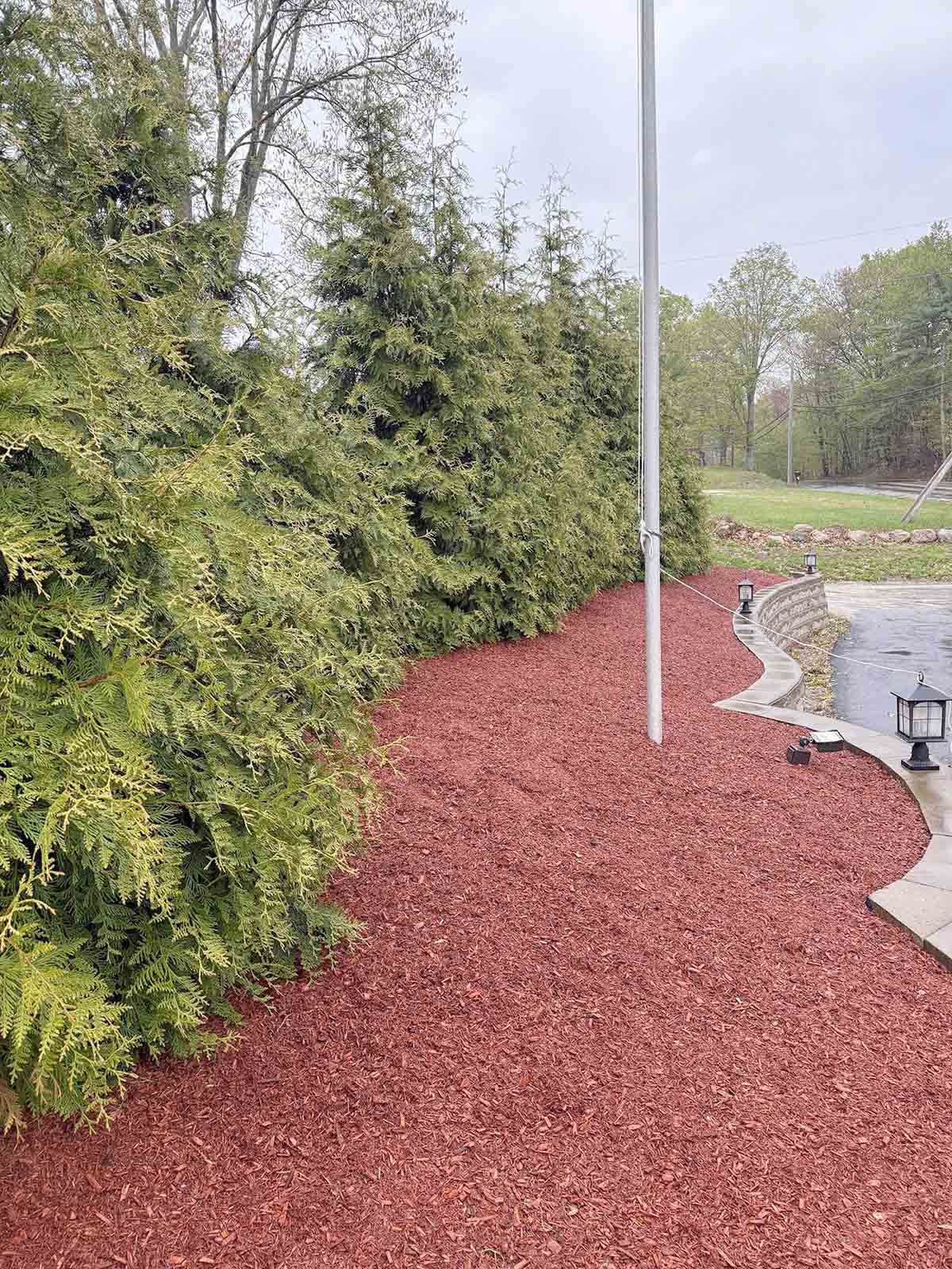 Red gravel path beside dense green shrubs and a stone patio with a flagpole.
