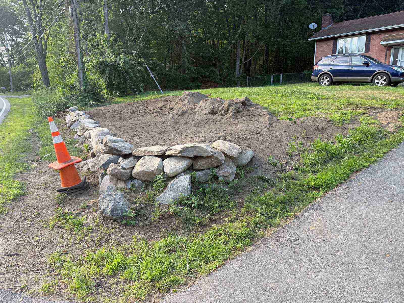 Stone-bordered dirt patch beside a road with orange cones, grass, trees, and a parked car nearby.