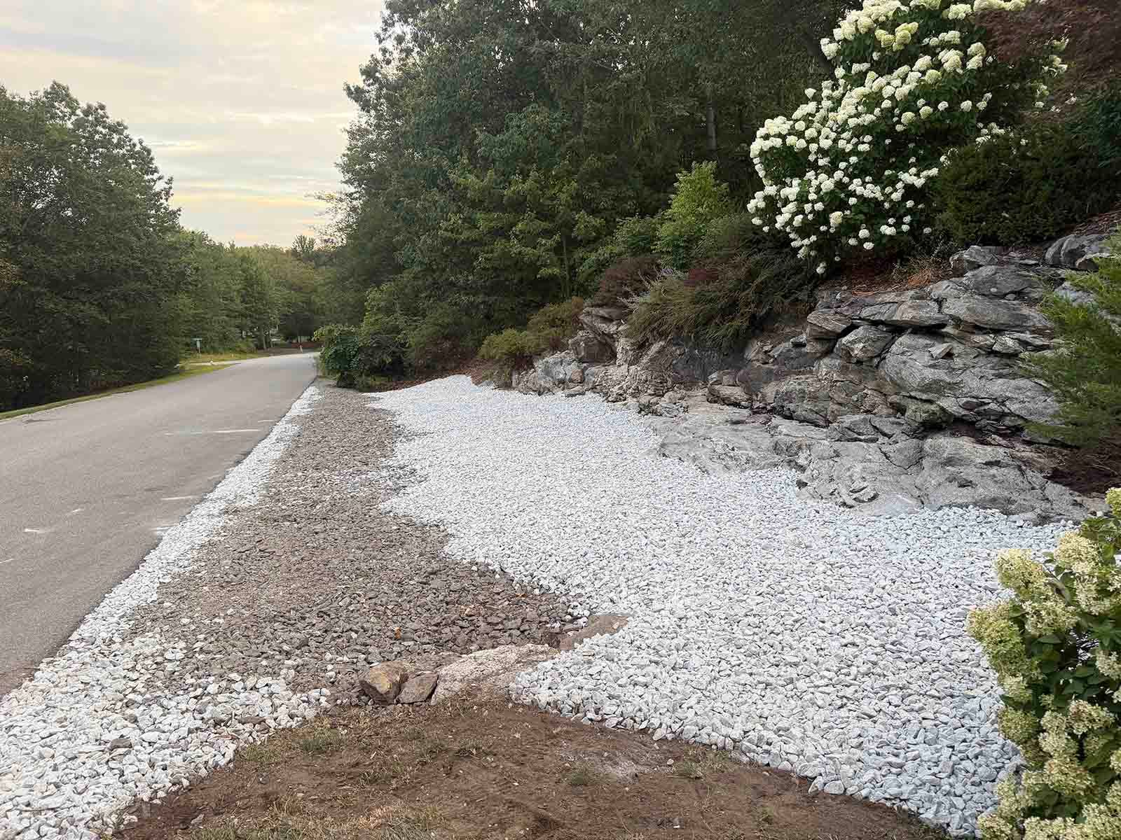 Gravel shoulder beside a road, with white rock landscaping and trees along a rocky embankment.