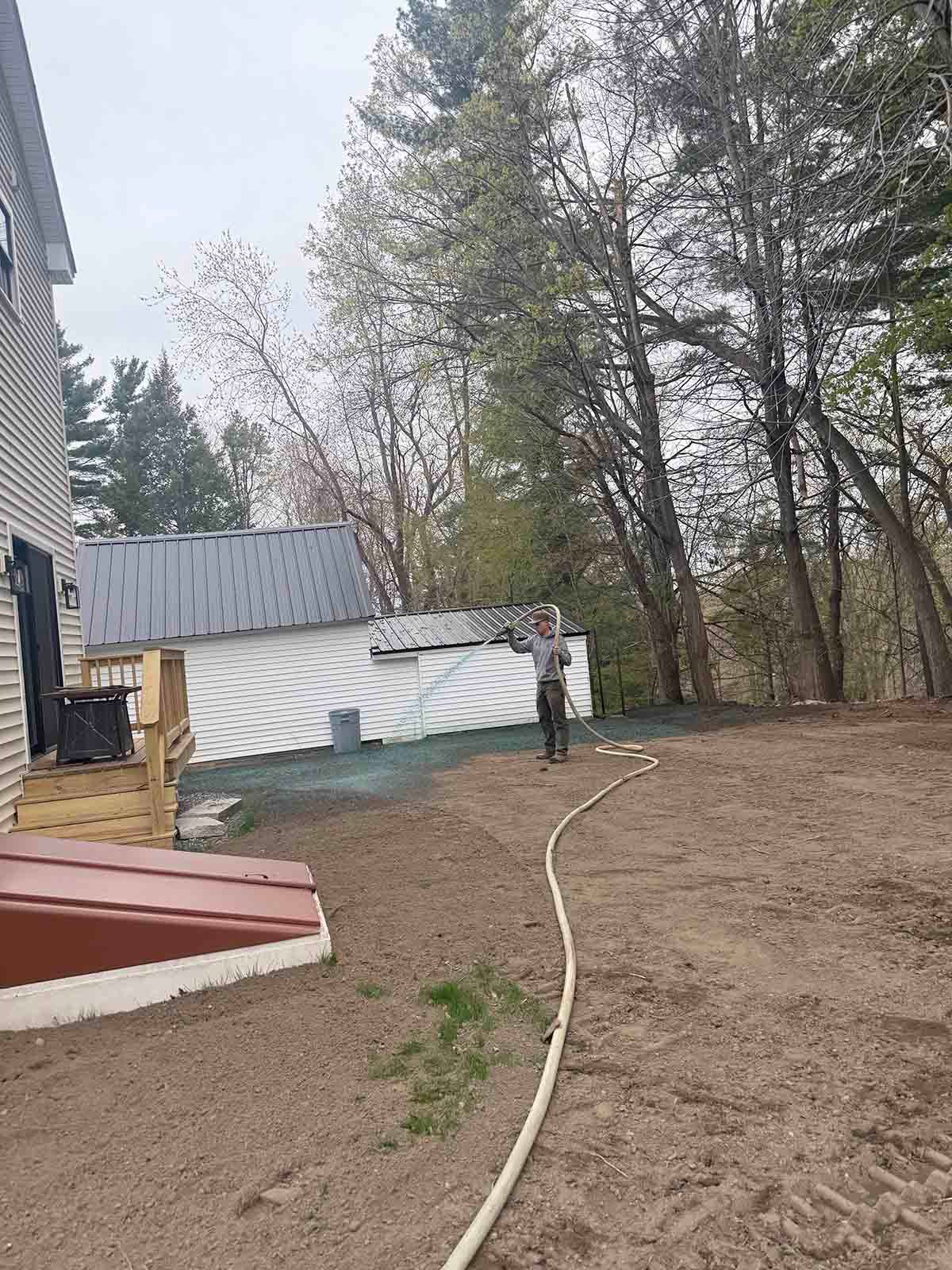 Backyard construction site with a shed, dirt ground, and a hose leading toward trees