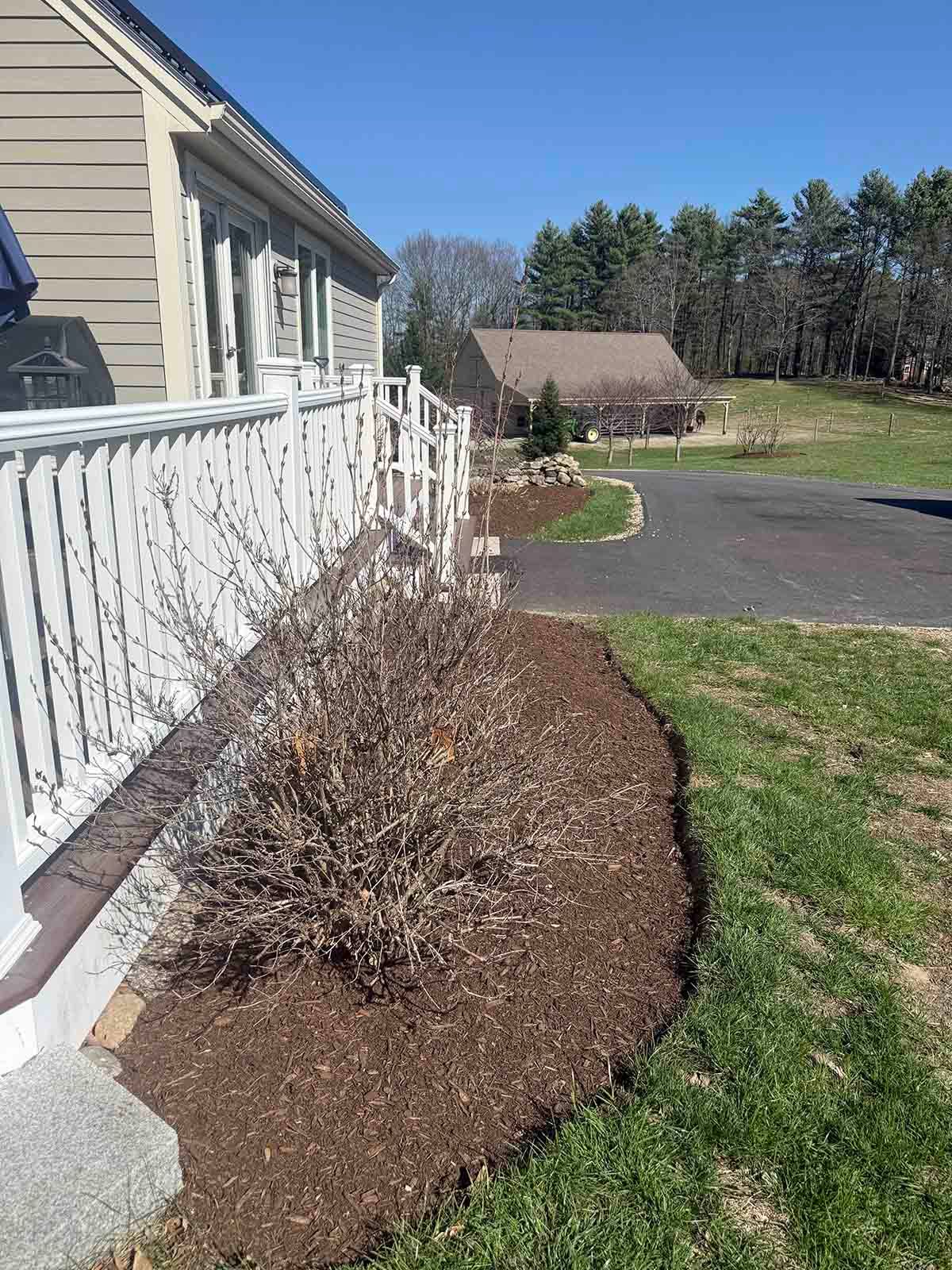 Front yard with white porch railing, mulched shrub bed, green lawn, and driveway under a clear sky