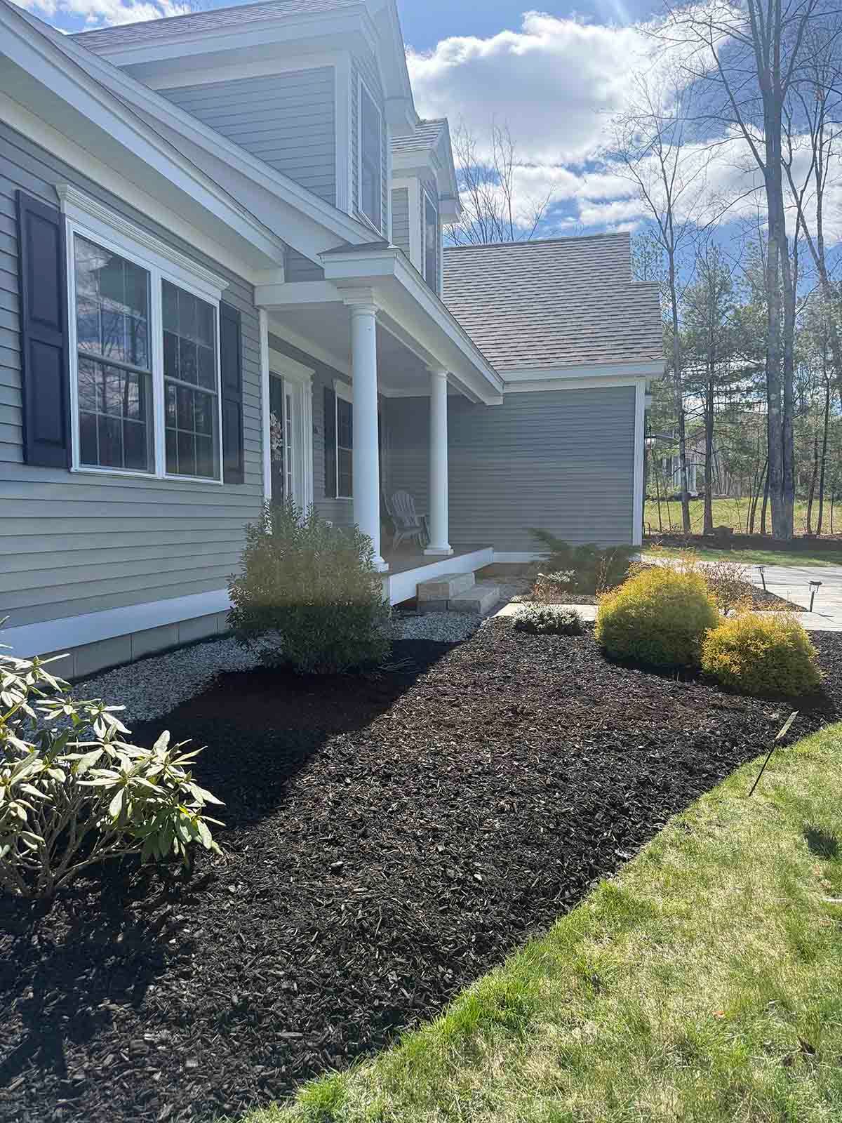 Front of a gray house with white trim, shrubs, mulch bed, and green lawn under a blue sky
