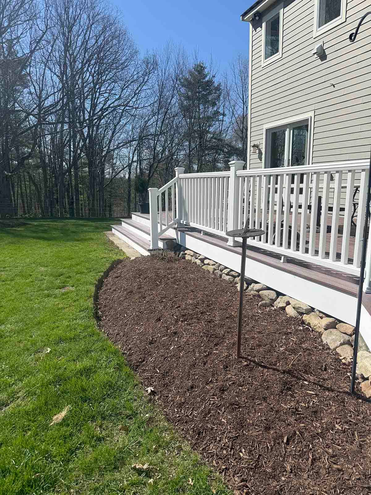House deck with white railing beside a mulched garden bed and grassy yard under a clear sky
