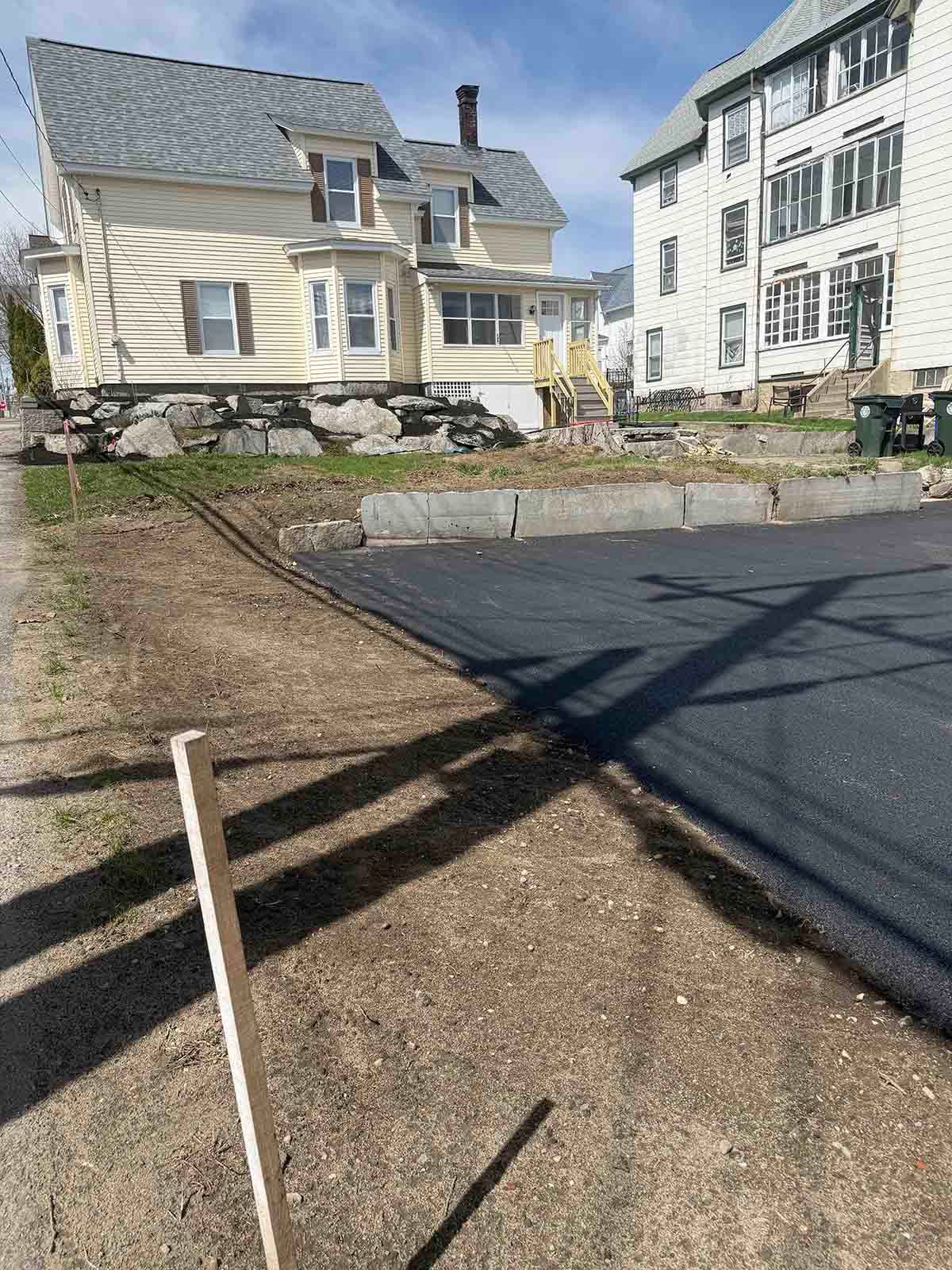 Residential buildings beside a freshly paved driveway with a small lawn and stone curb under sunny skies