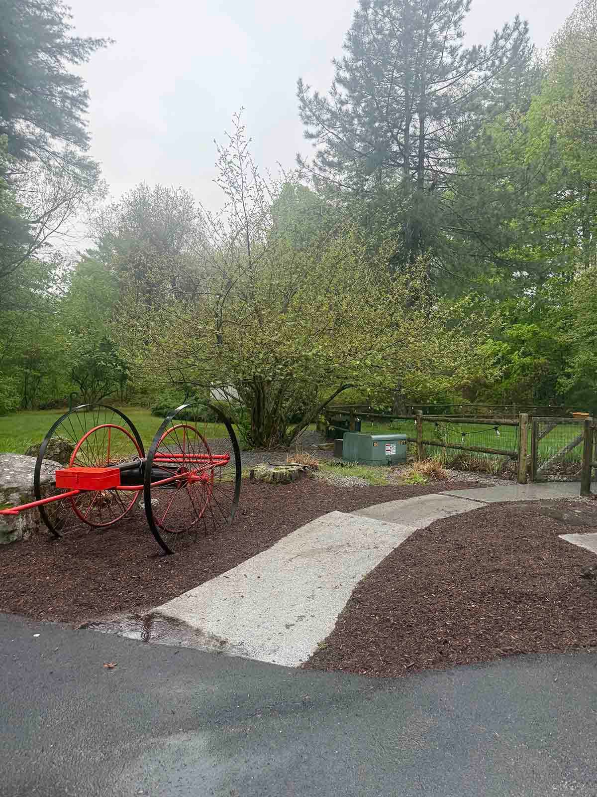 Park with a red farm-style play wheel, curved gravel path, and trees on a wet day